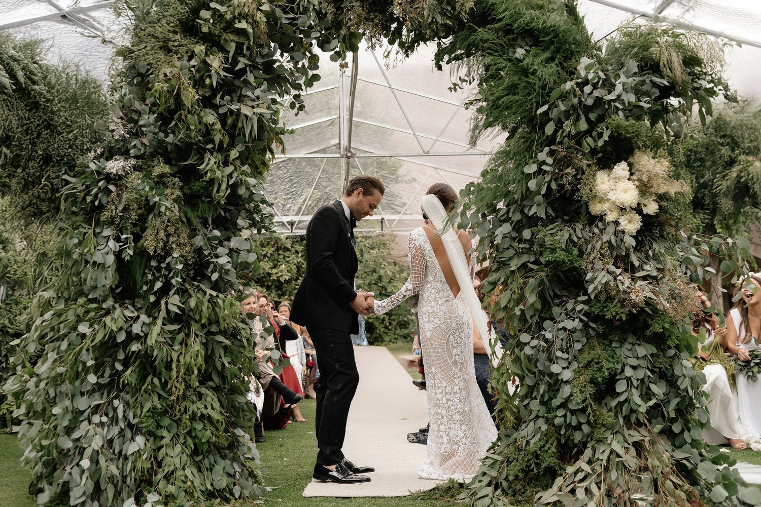 A couple stands beneath a lush green floral arch at an elegant garden wedding ceremony.