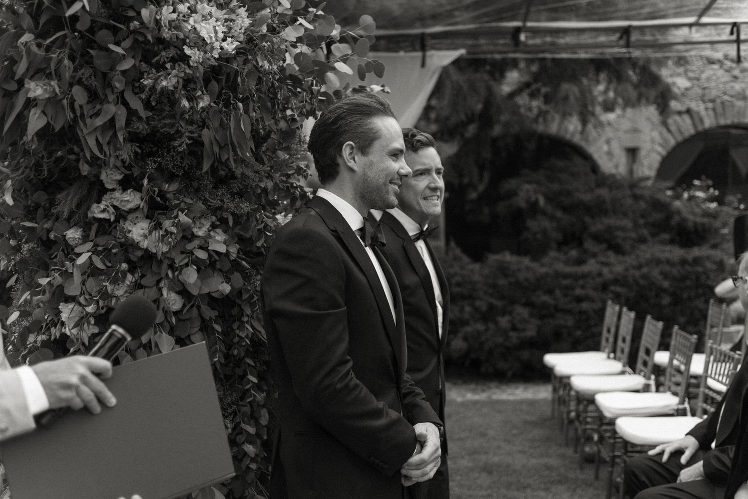 Two men in formal black suits stand at attention during an outdoor ceremony with white chairs arranged in rows.