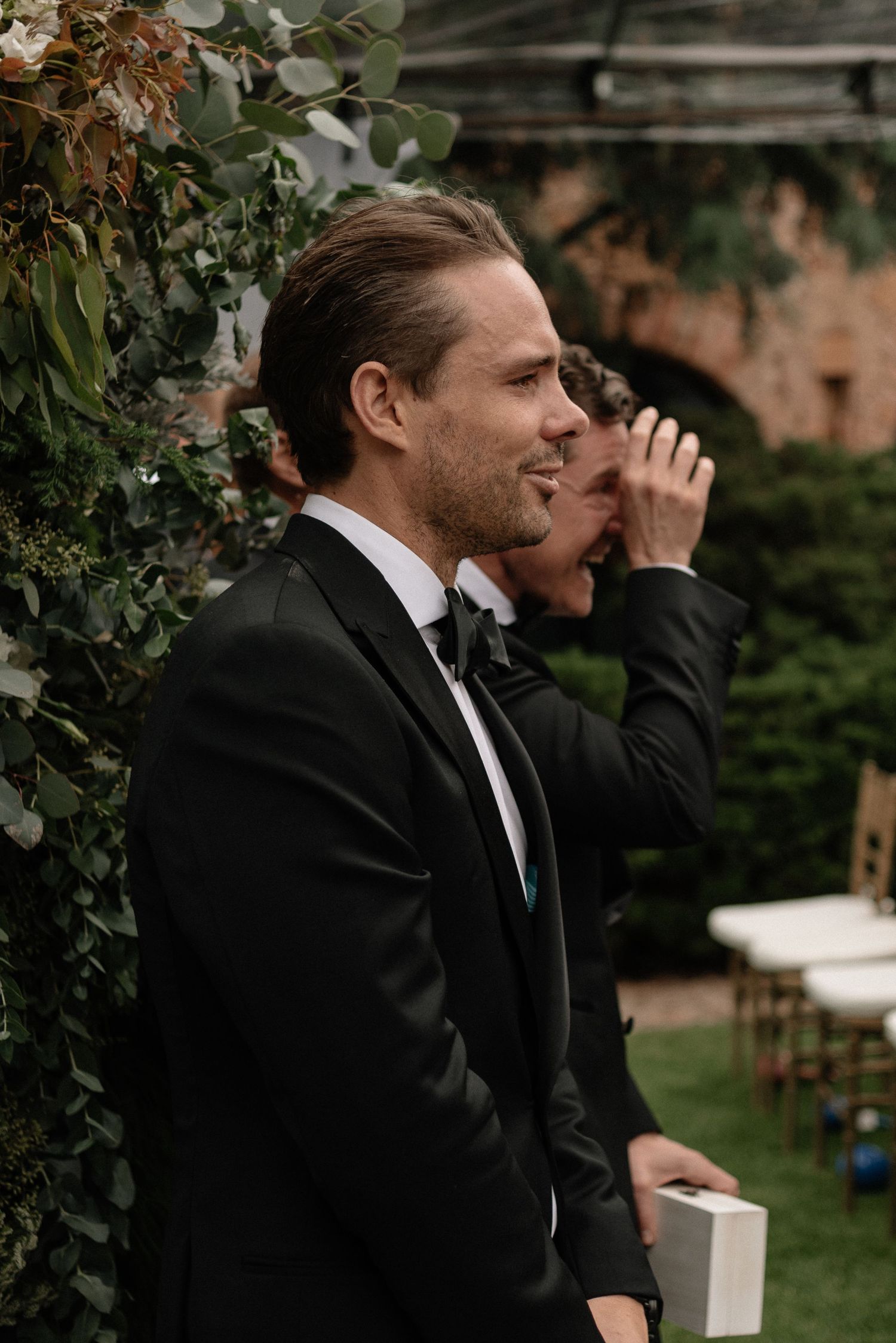 A groom in a classic black tuxedo and bowtie stands thoughtfully by lush garden foliage at an outdoor wedding venue.
