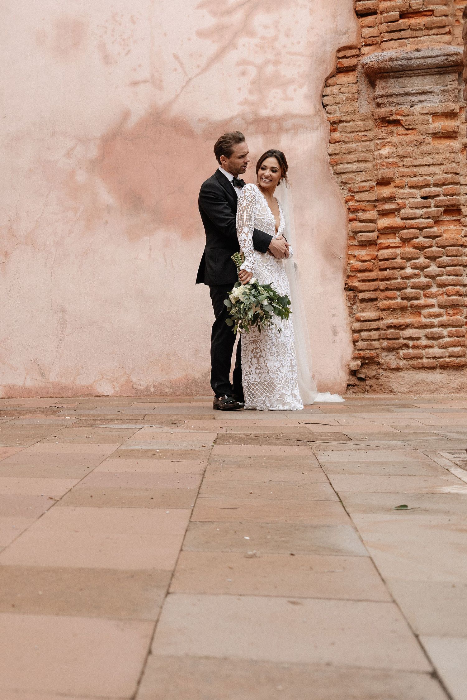 A bride and groom pose together against a rustic brick wall on their wedding day, with the bride holding a floral bouquet.