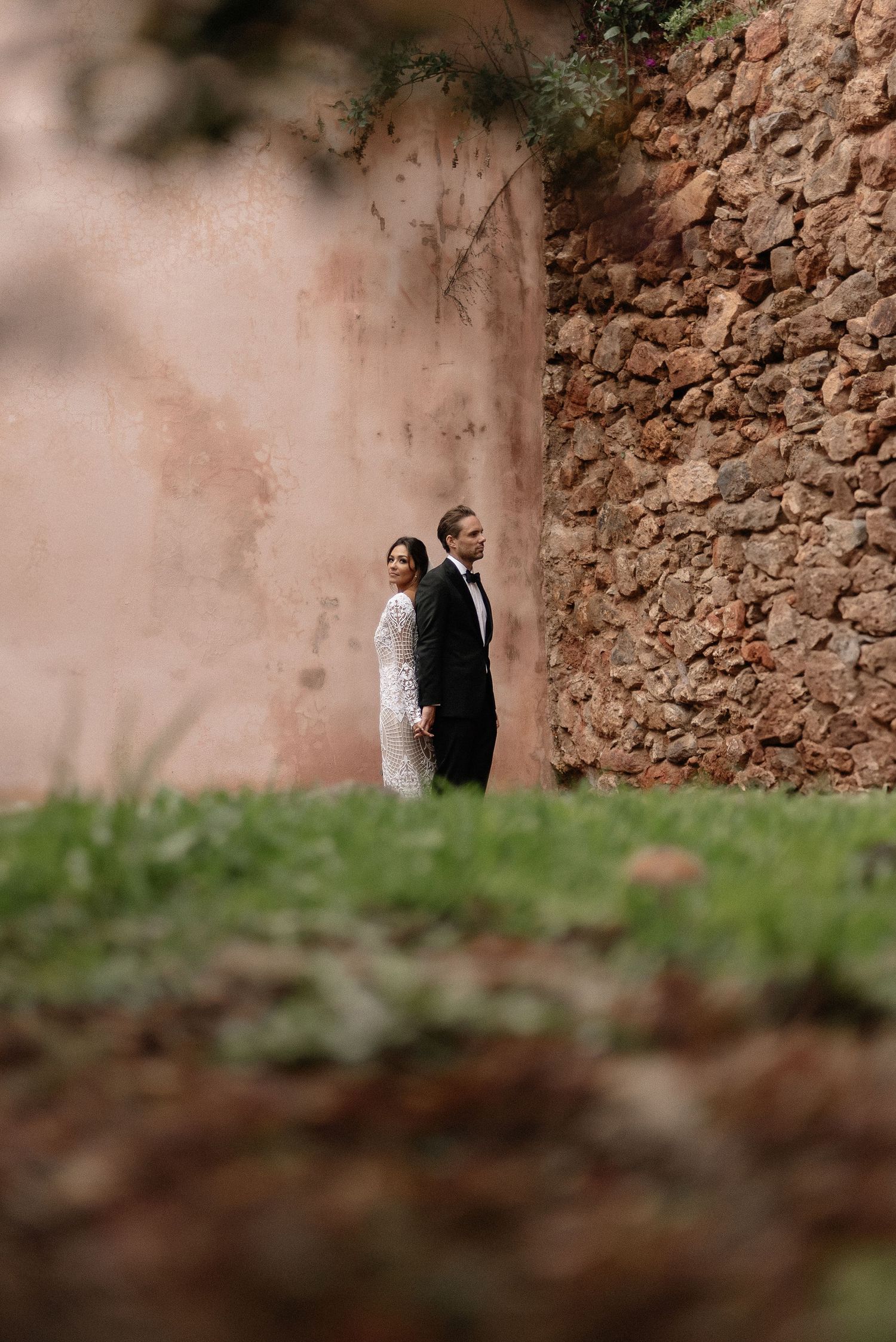 A couple in wedding attire stands against a rustic stone wall in a romantic outdoor setting.