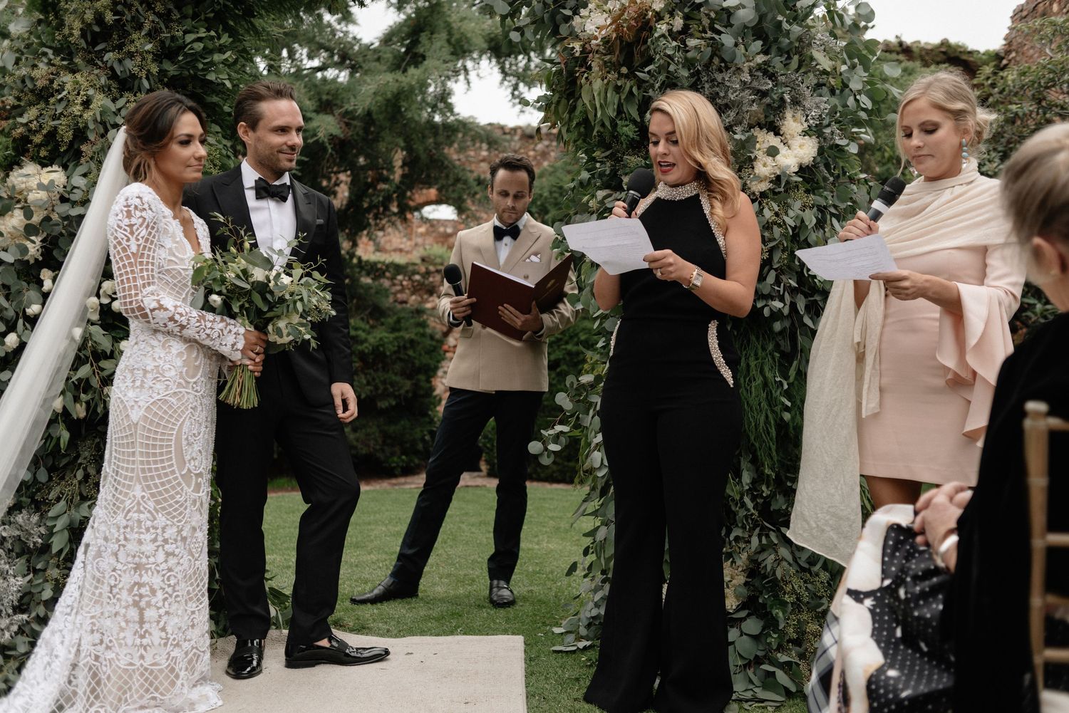 Wedding ceremony takes place in a garden setting with flowers and greenery surrounding the attendees and officiant.