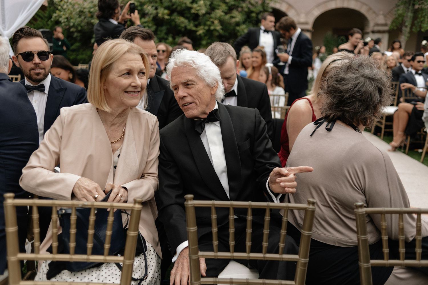 Elegantly dressed party guests sit in tuxedos and formal attire at an outdoor social gathering with arched architecture.