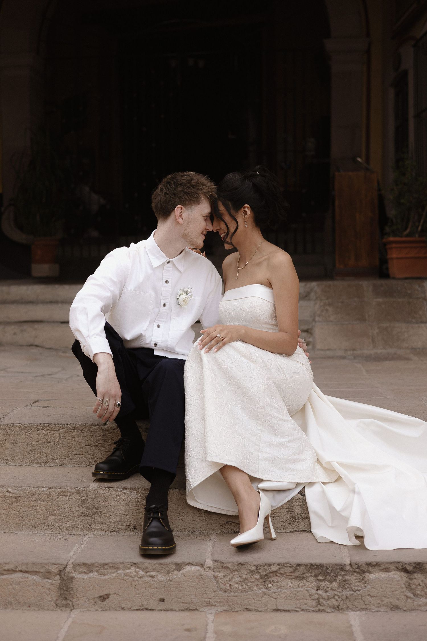 A romantic wedding couple sits together on stone steps, dressed in formal white attire and sharing an intimate moment.