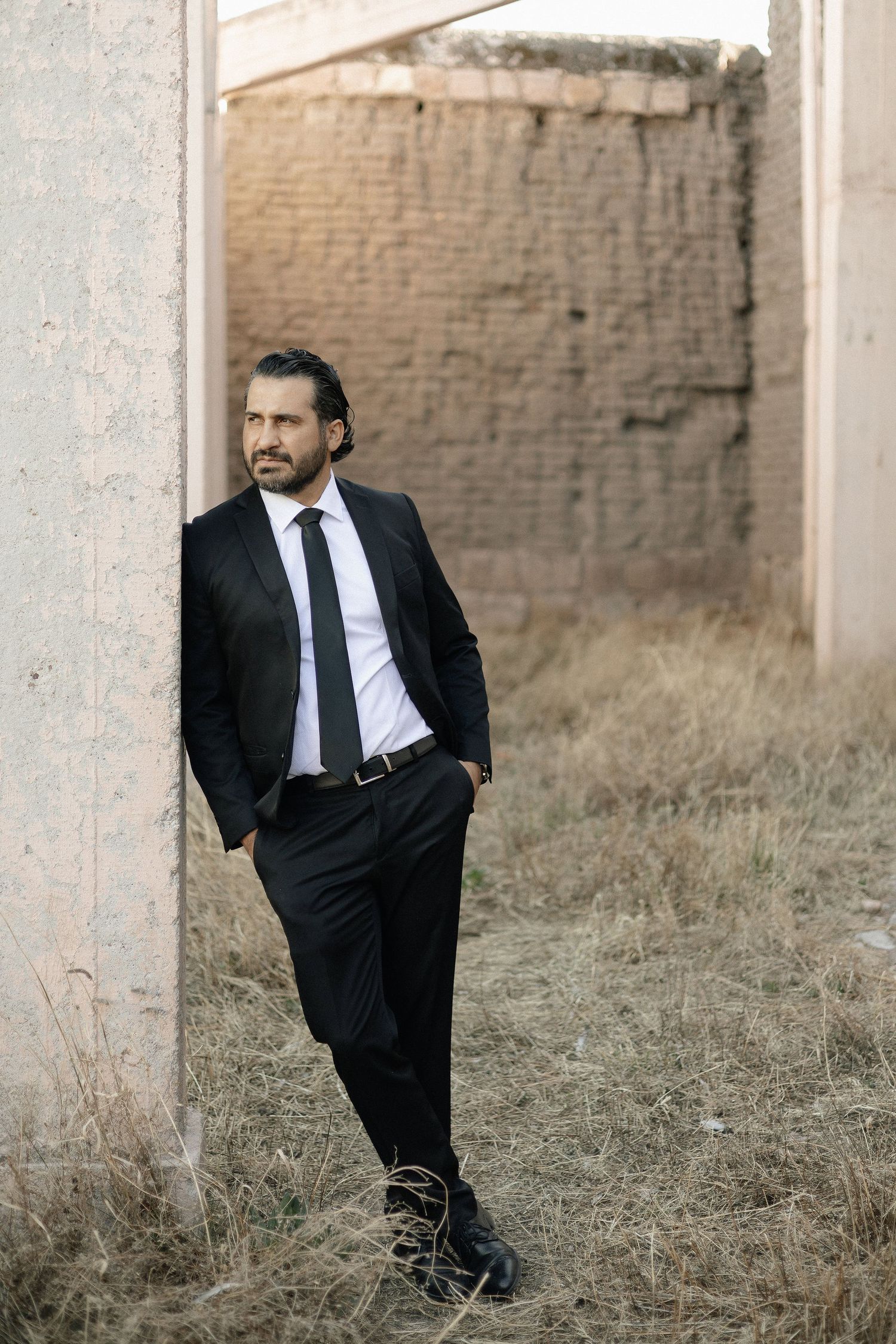A person in a black suit stands in tall dry grass against a brick wall background.