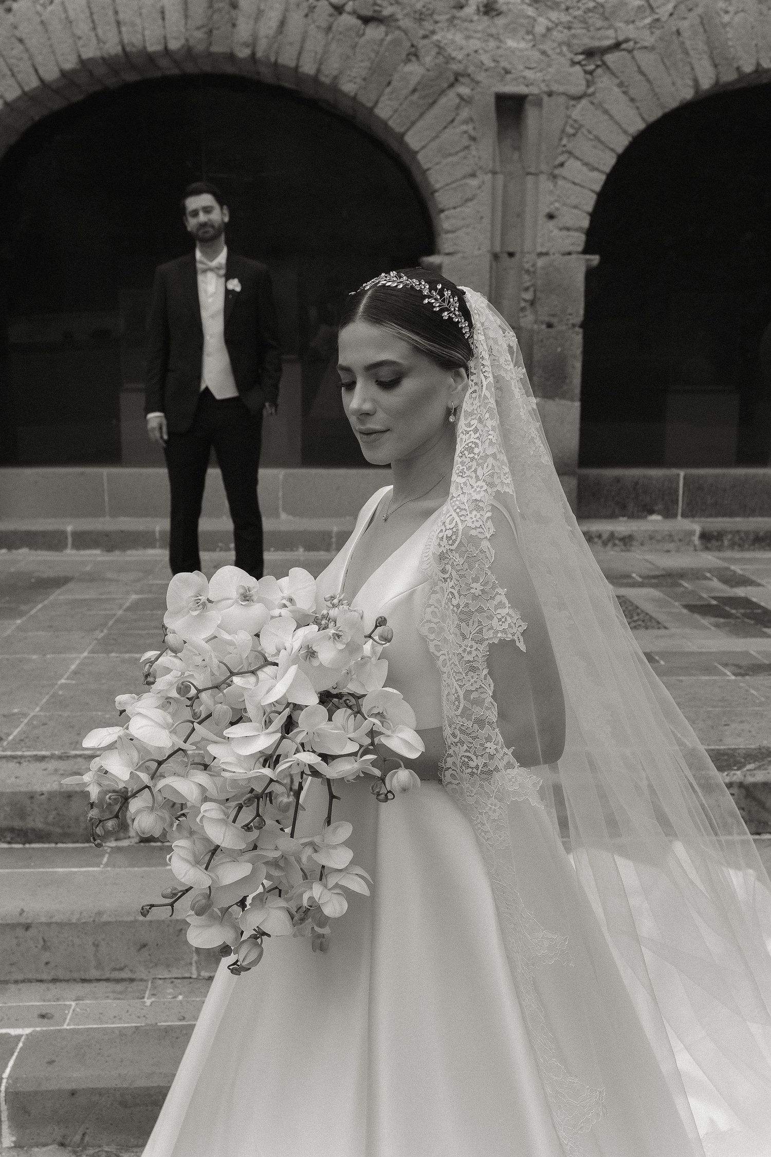 Black and white wedding photo showing a bride in an elegant gown with veil and bouquet near stone archways.