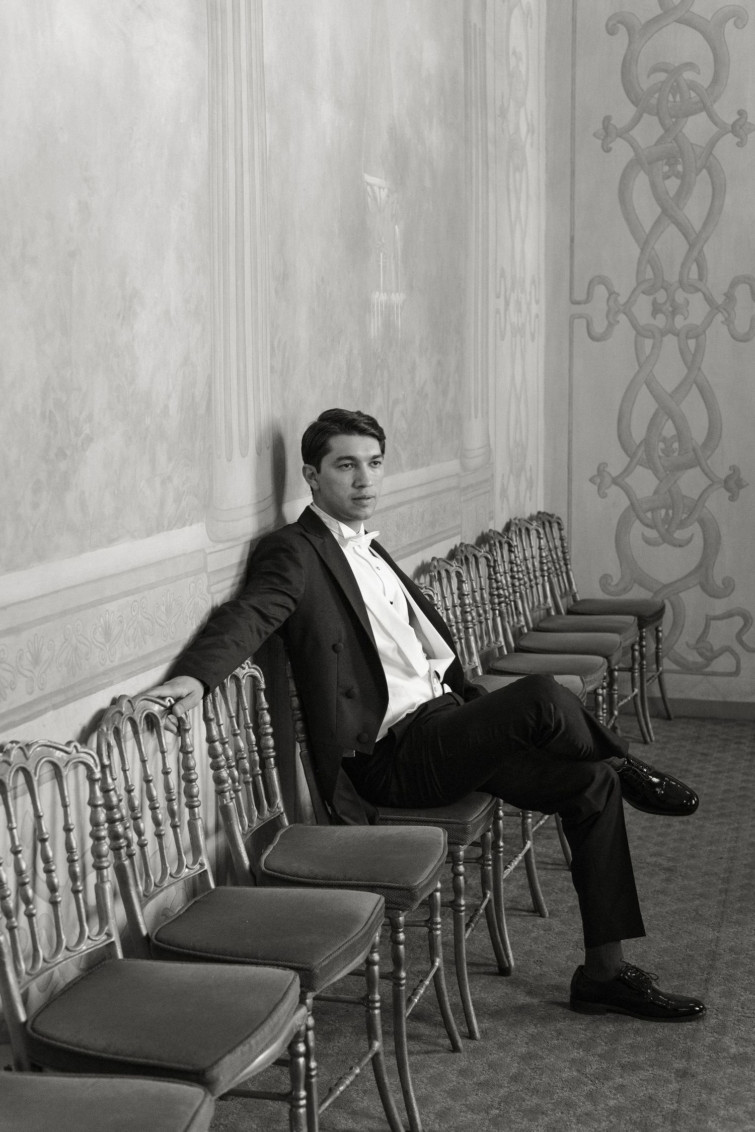 A black and white photograph shows a person sitting alone in a row of ornate chairs against a patterned wall.