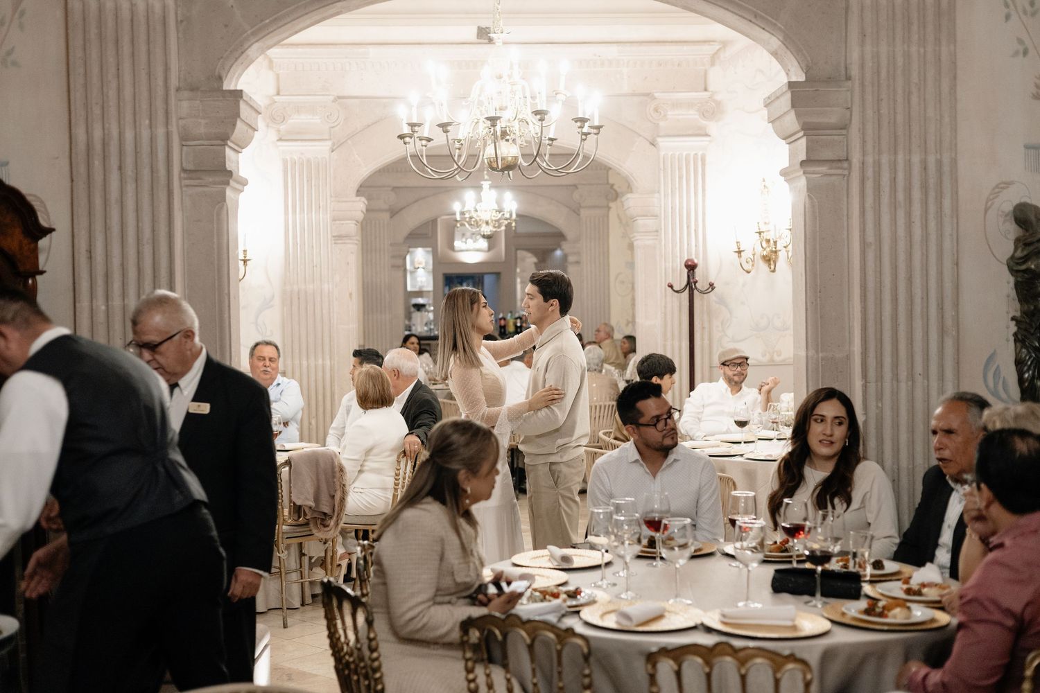 Elegant wedding reception in a white ballroom with crystal chandeliers and guests seated at formal dining tables.