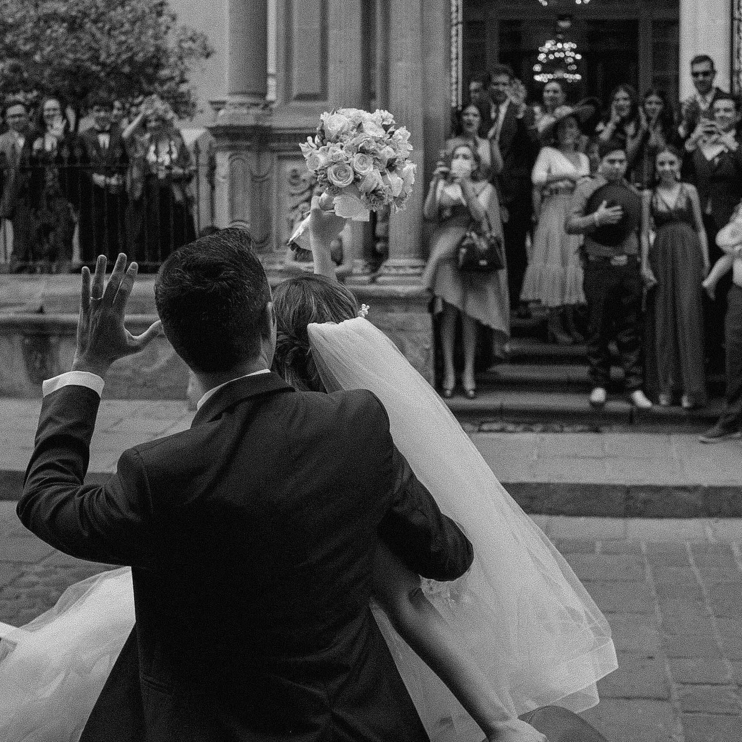 A newlywed couple waves to guests gathered on church steps in this black and white wedding photograph.