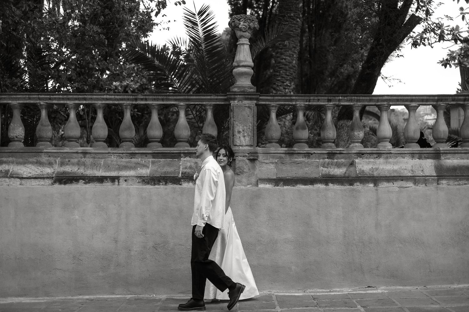 A romantic wedding couple walks along a stone balustrade in a garden setting during their outdoor photoshoot.