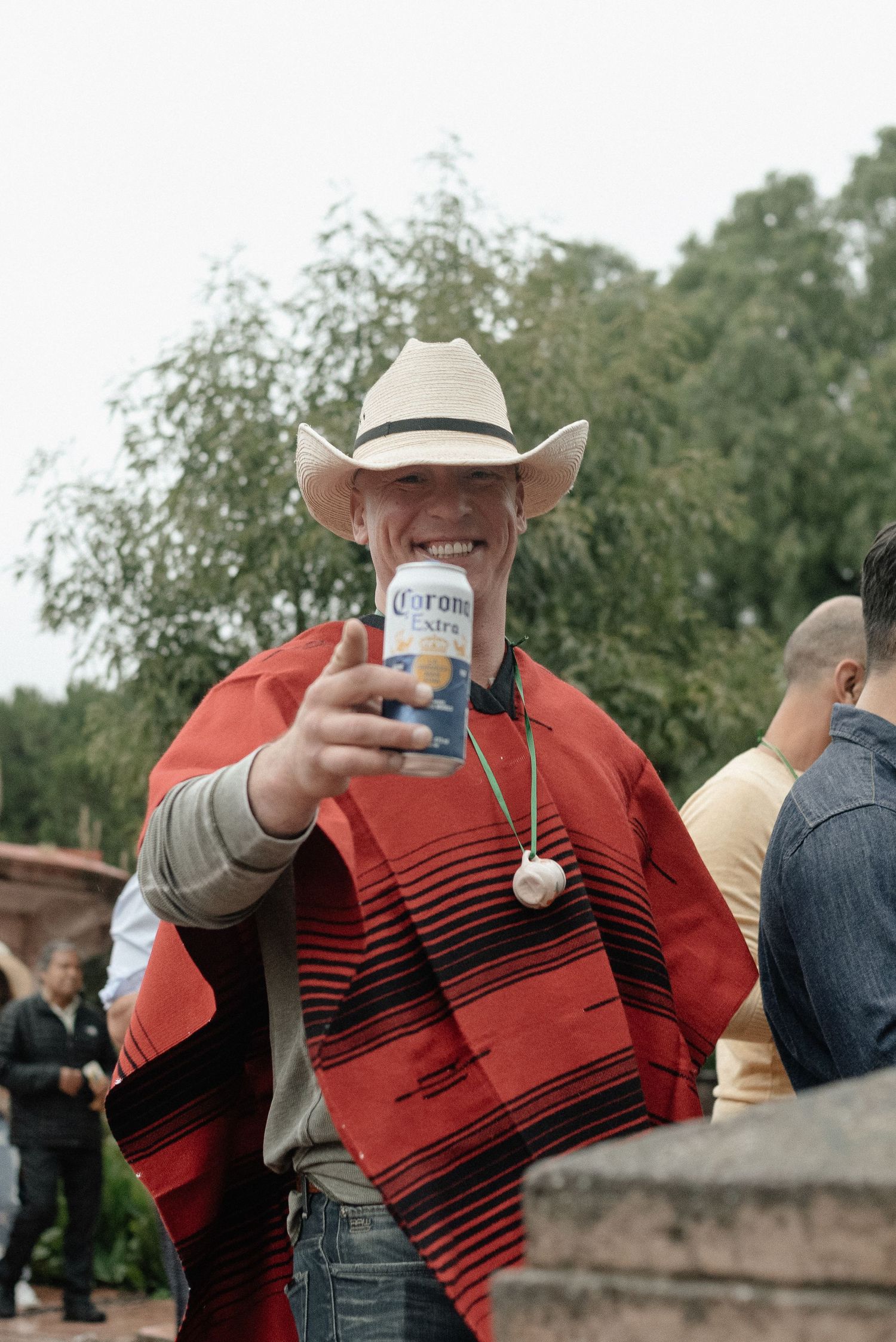 A person in a red striped poncho and cowboy hat holds up a Corona beer while celebrating at an outdoor event.