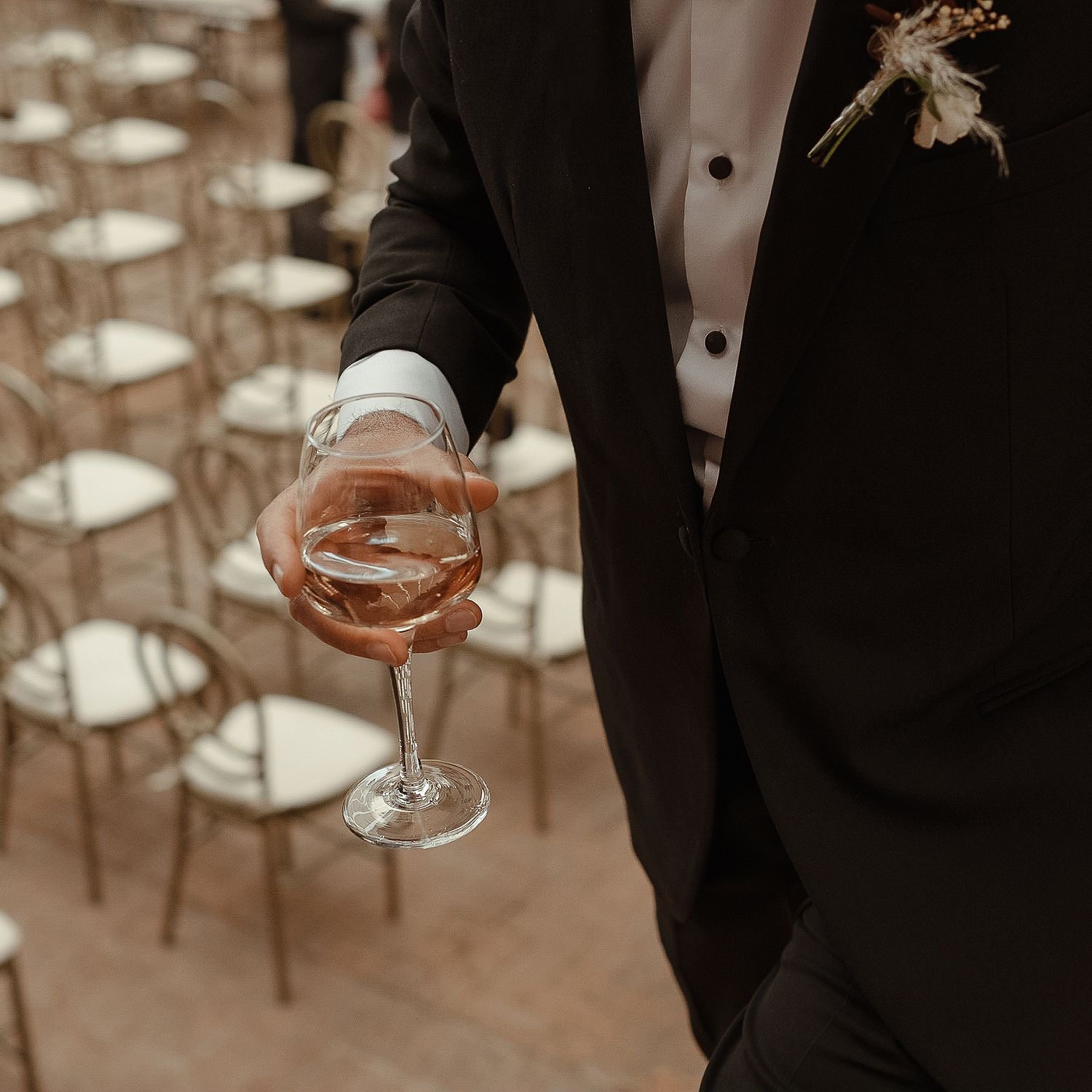 A suited arm holding a glass of rosé wine with empty chairs in the background.