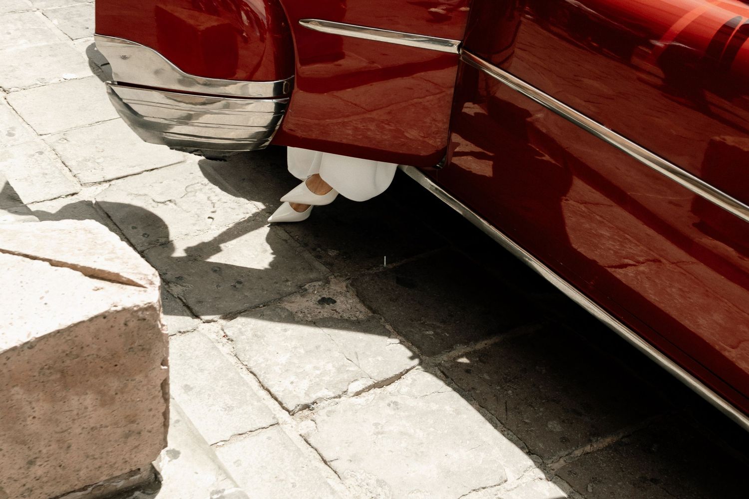 A vintage red car's chrome bumper and fender casting shadows on cracked concrete pavement.