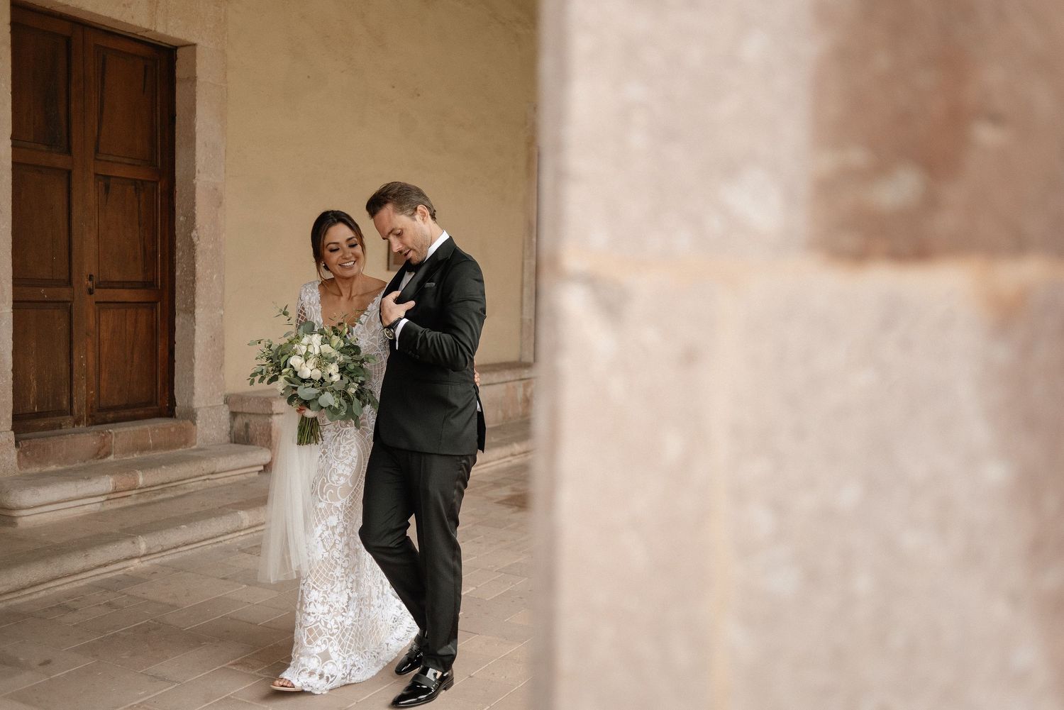 Two people in wedding attire share an intimate moment against a neutral-toned wall with wooden door detail.