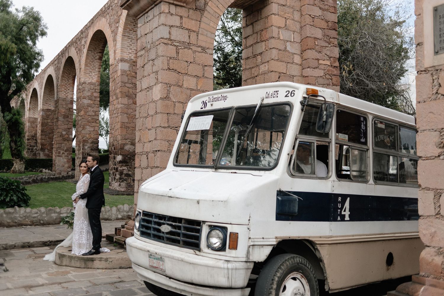 A vintage white shuttle bus parked in front of a historic stone archway and ivy-covered wall.