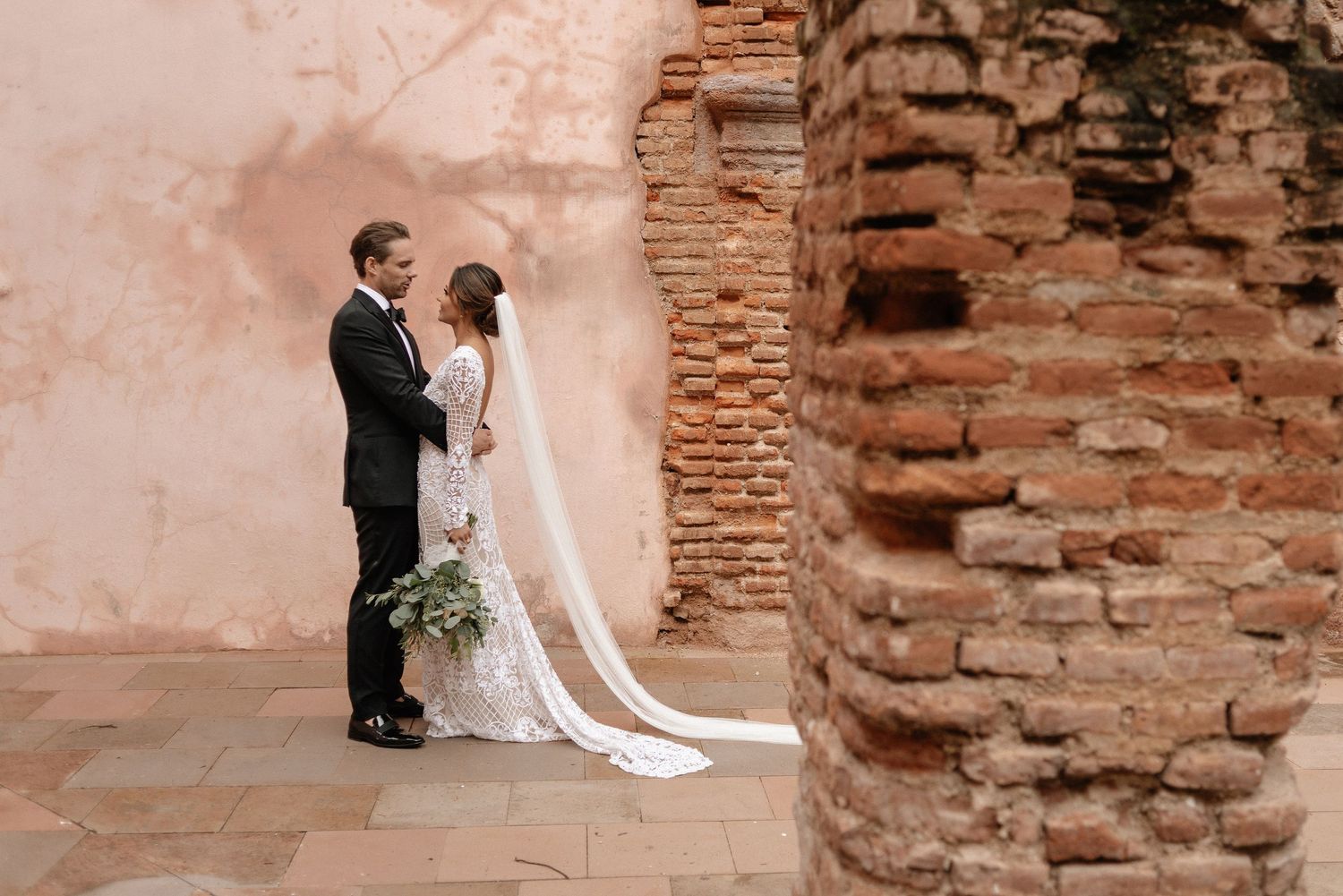 Wedding couple stands beside rustic brick wall during romantic portrait session at historic venue.