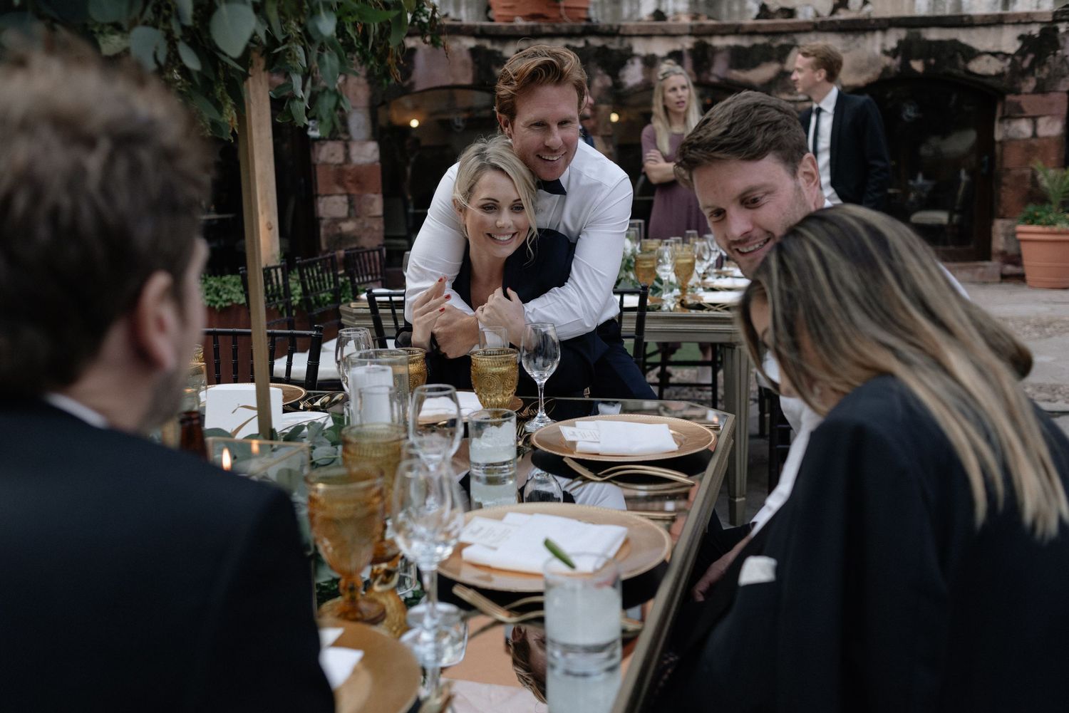A waiter serves a group of restaurant patrons seated at an outdoor dining table with drinks and place settings.