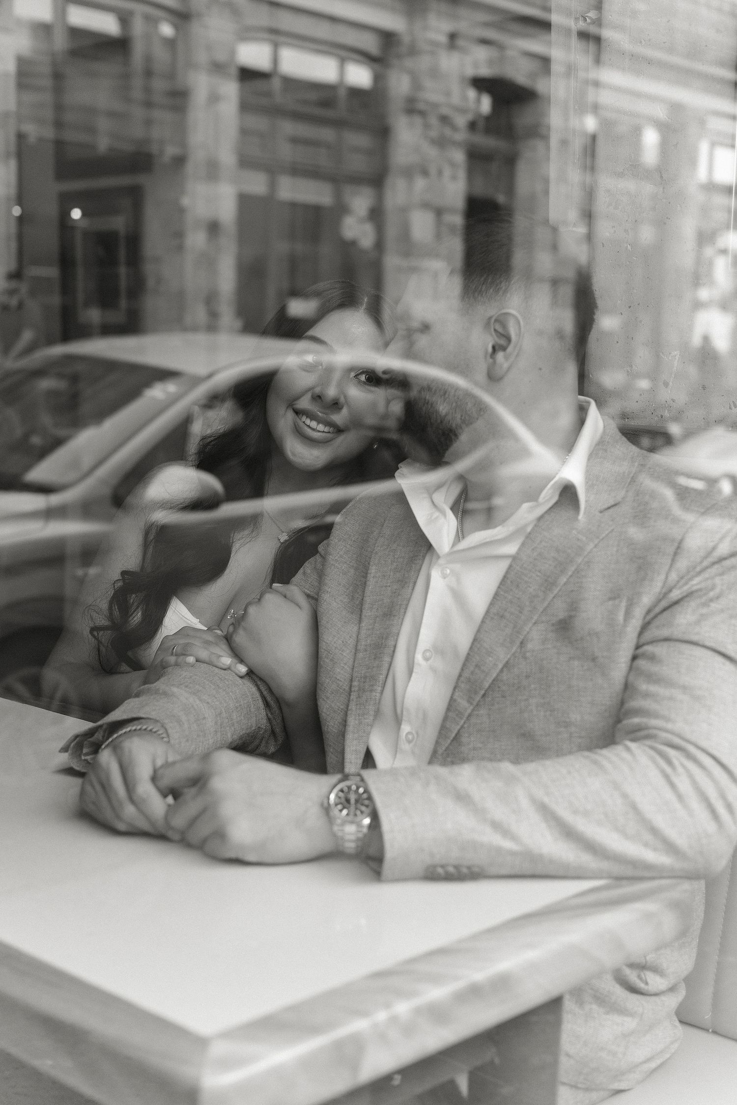 A black and white photograph showing a couple sitting at a table, captured through a window reflection.