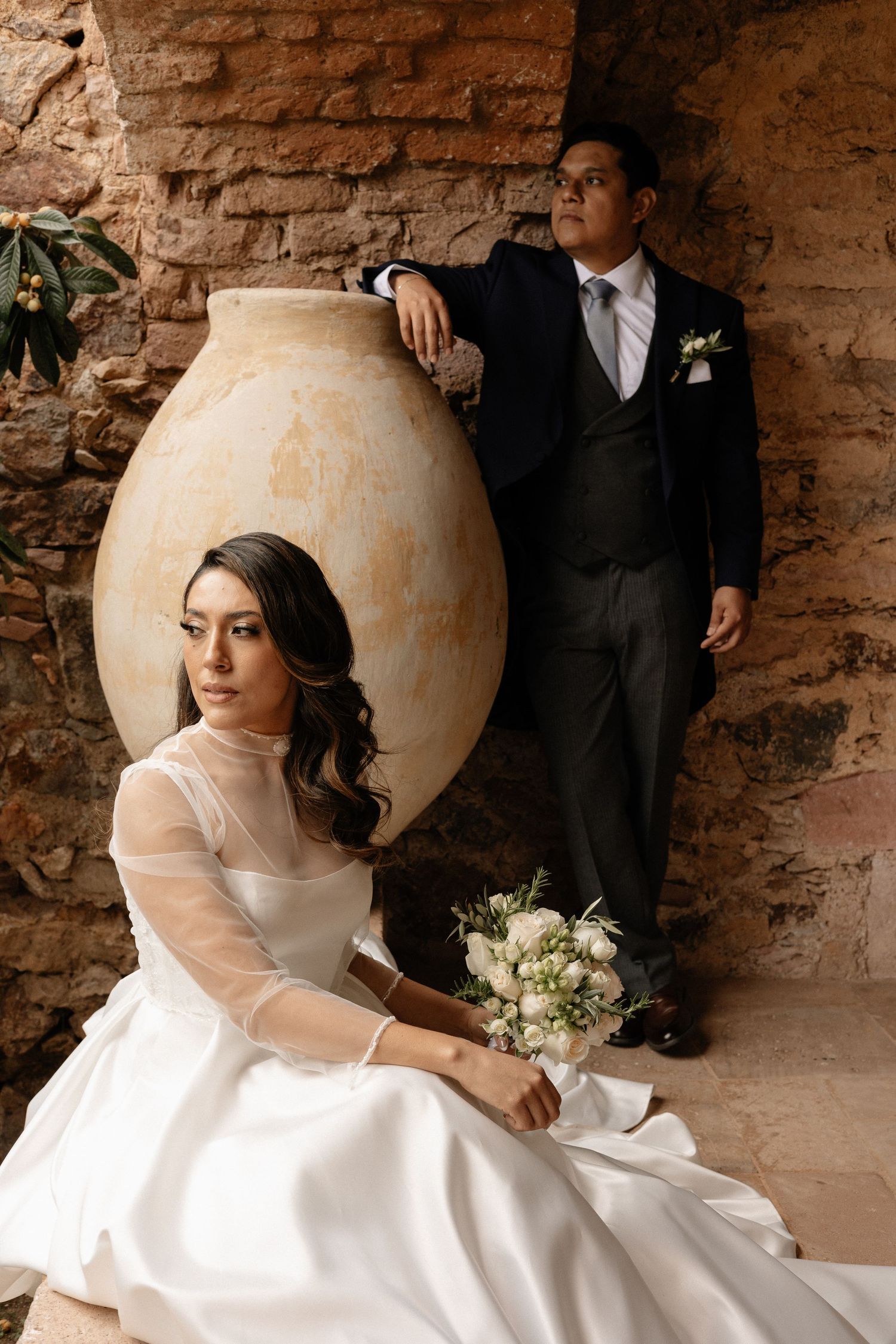 A wedding couple poses artistically with a large clay amphora against rustic stone walls.