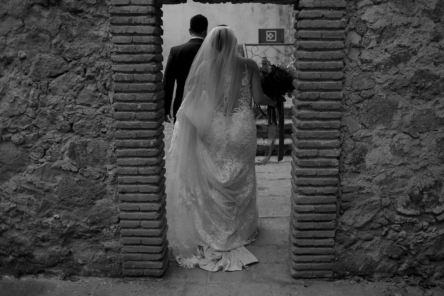 Black and white photo of a bride and groom walking through a stone archway.
