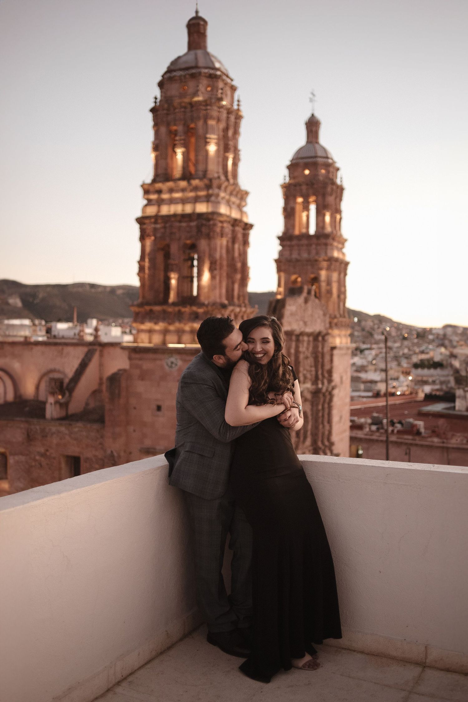 Romantic couple embracing on a rooftop with historic Mexican cathedral towers at sunset.