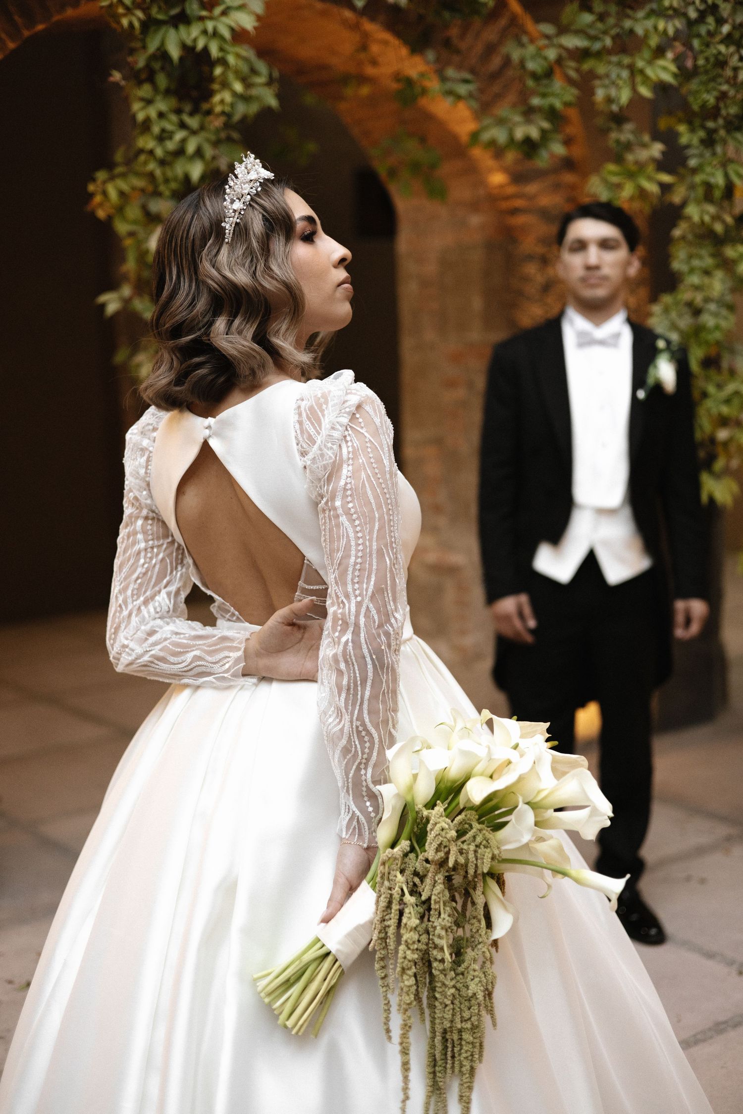 Elegant wedding photo featuring a bride in a lace-sleeved gown holding white flowers while groom stands in background.