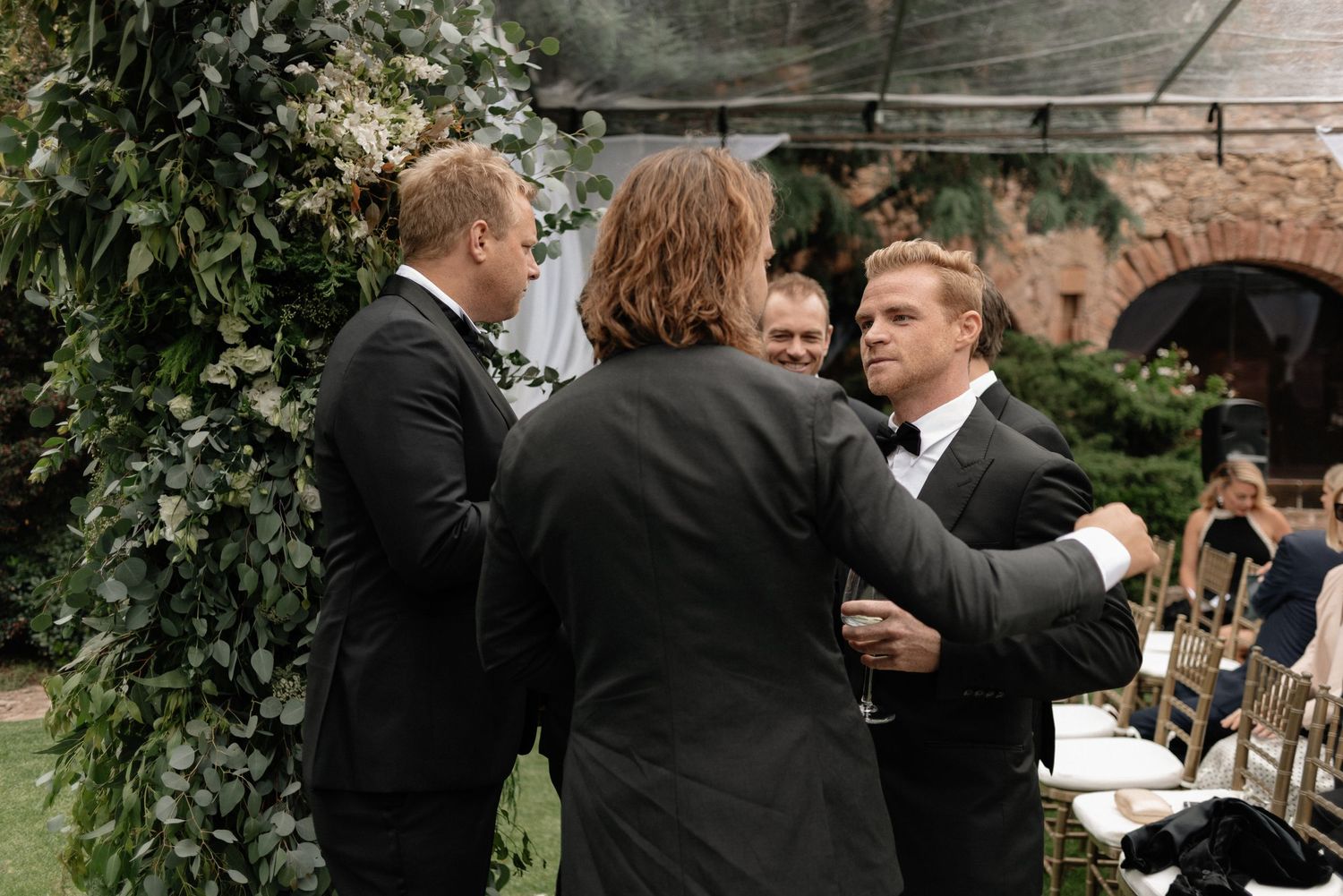 Three people dressed in black suits having an animated conversation at an outdoor event with greenery and seating in background.