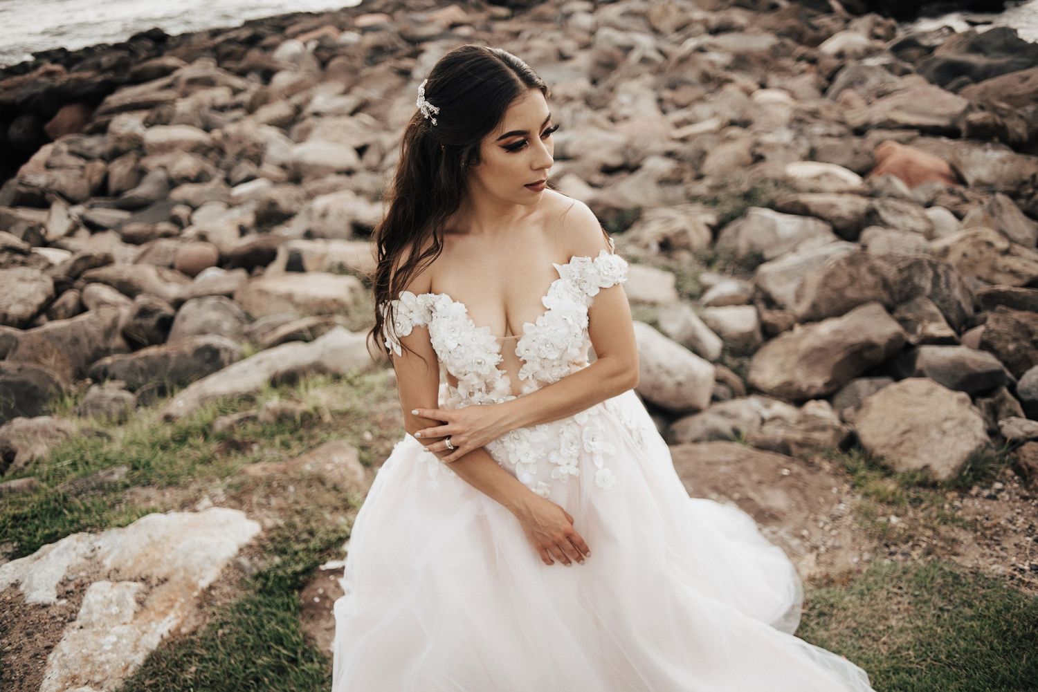 A bride in a flowing white dress poses against a rustic stone wall backdrop.