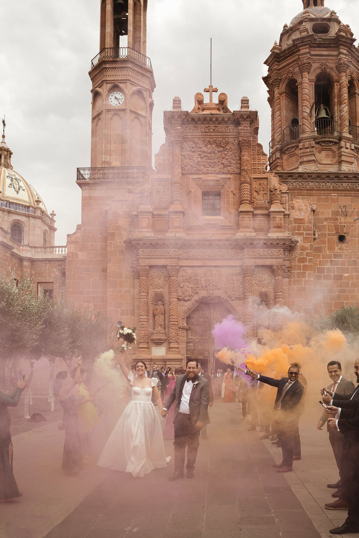 Colorful smoke fills the air outside a historic church during a wedding celebration.