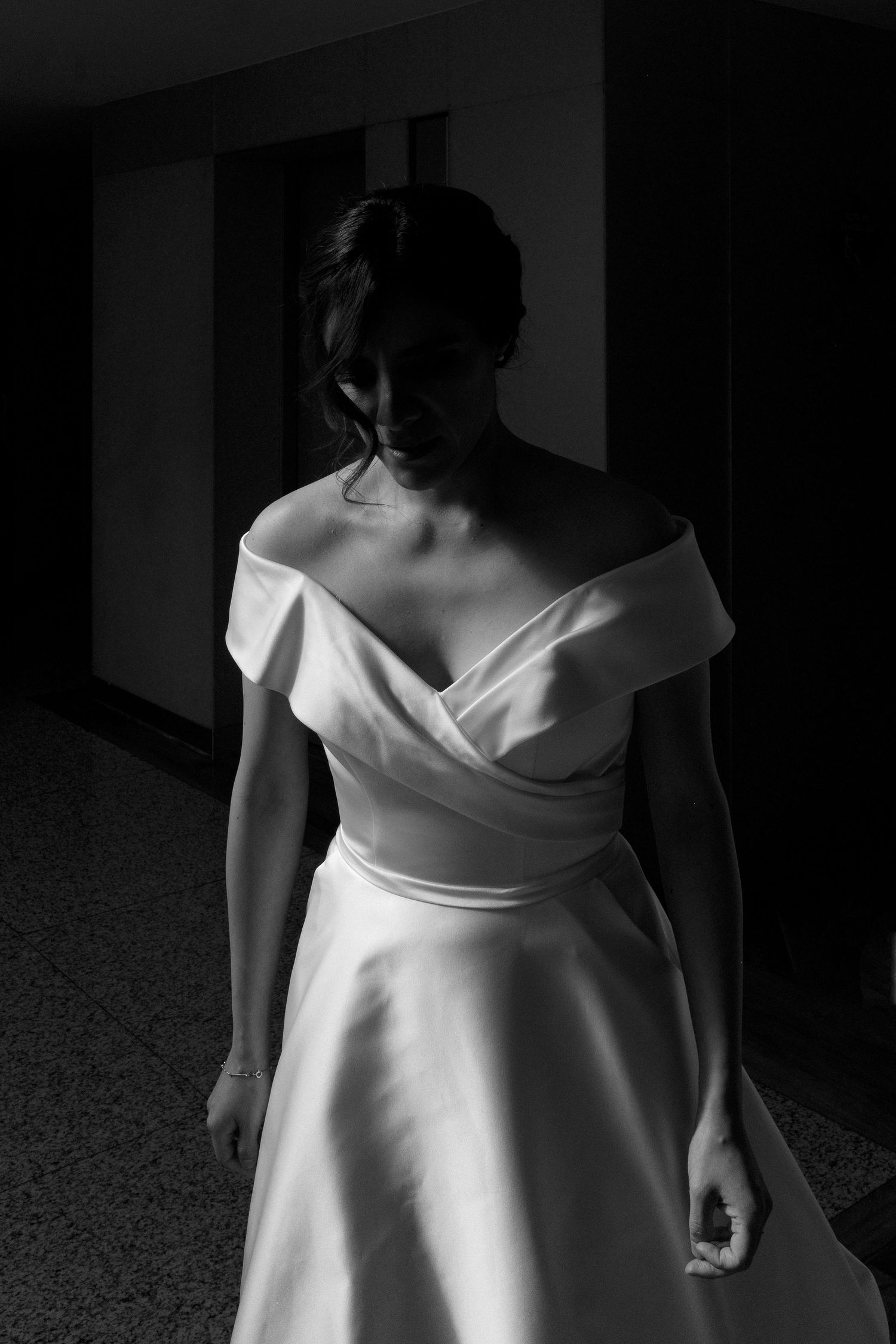 Dramatic black and white portrait of off-shoulder wedding gown in dark hallway.
