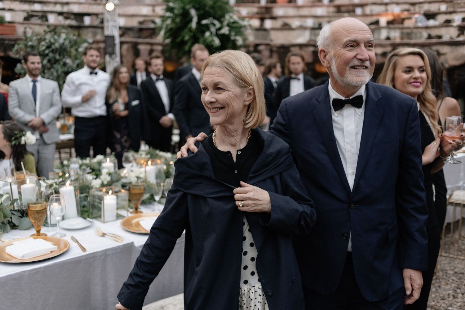Elegantly dressed couple laughs together at outdoor reception with table settings and guests in background.
