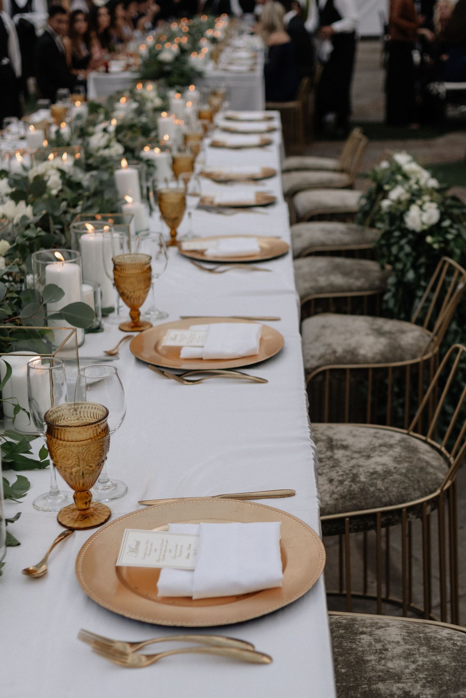 Elegant wedding table setting with amber goblets, gold chargers, white linens, and greenery floral arrangements.