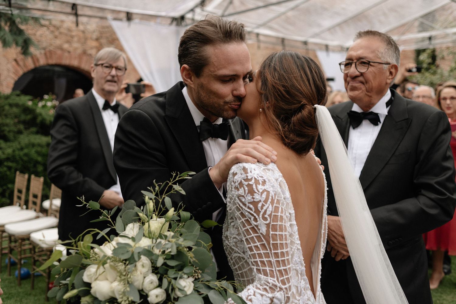 A wedding couple shares a kiss at an outdoor ceremony while holding a white floral bouquet.