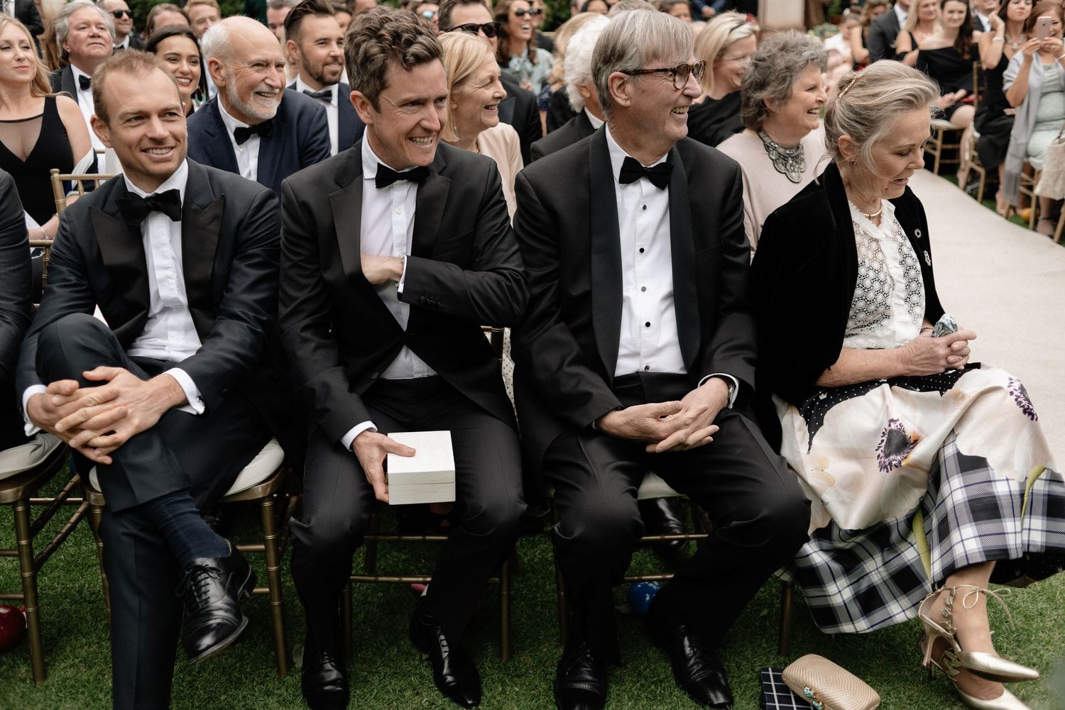 Wedding guests in formal attire seated in chairs outdoors during an elegant garden ceremony.