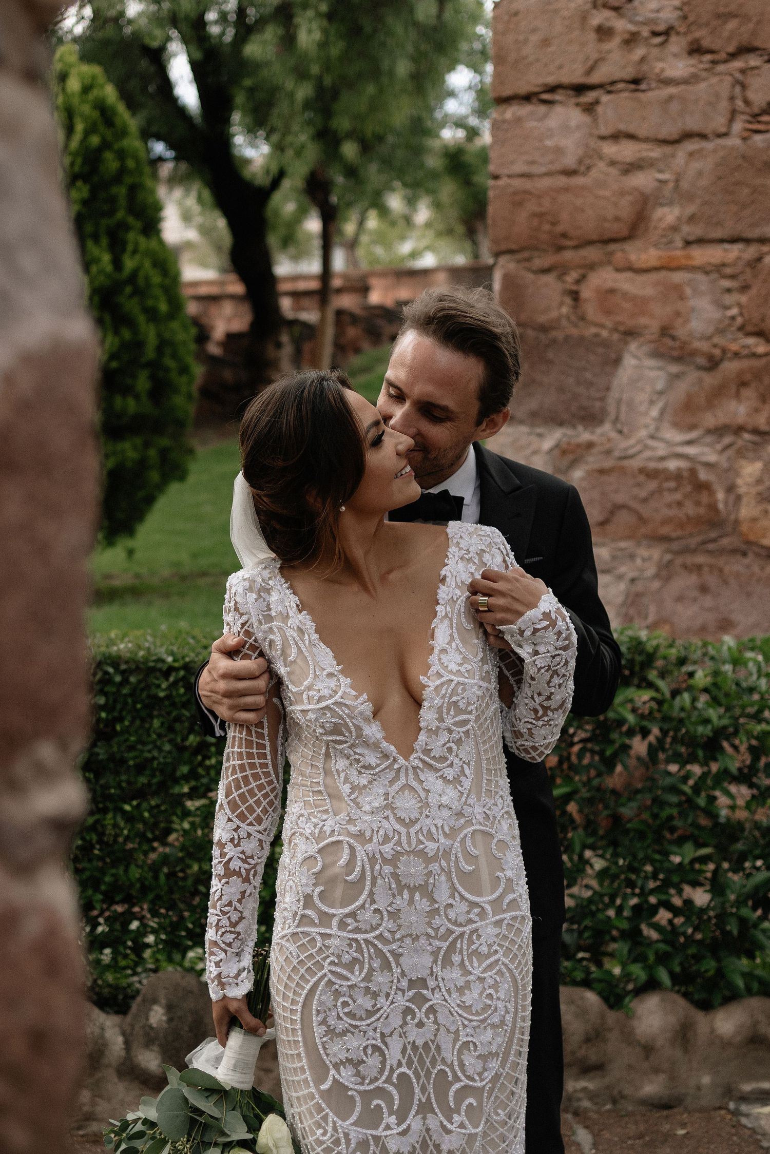 A romantic wedding couple embraces against a rustic stone wall backdrop, with the bride wearing an elegant lace gown.