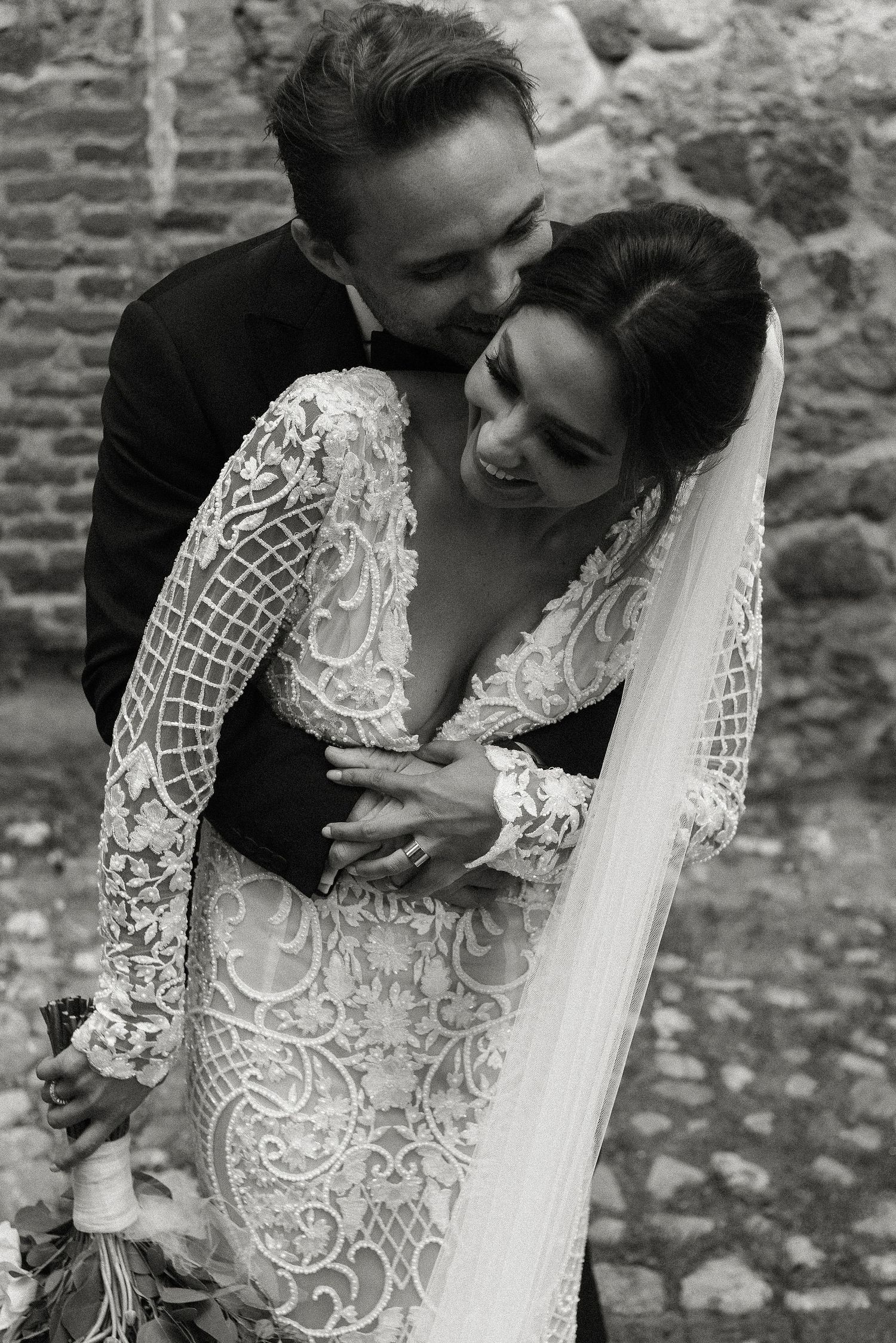 A romantic black and white wedding photo showing a tender embrace between newlyweds, with the bride in an intricate lace gown.