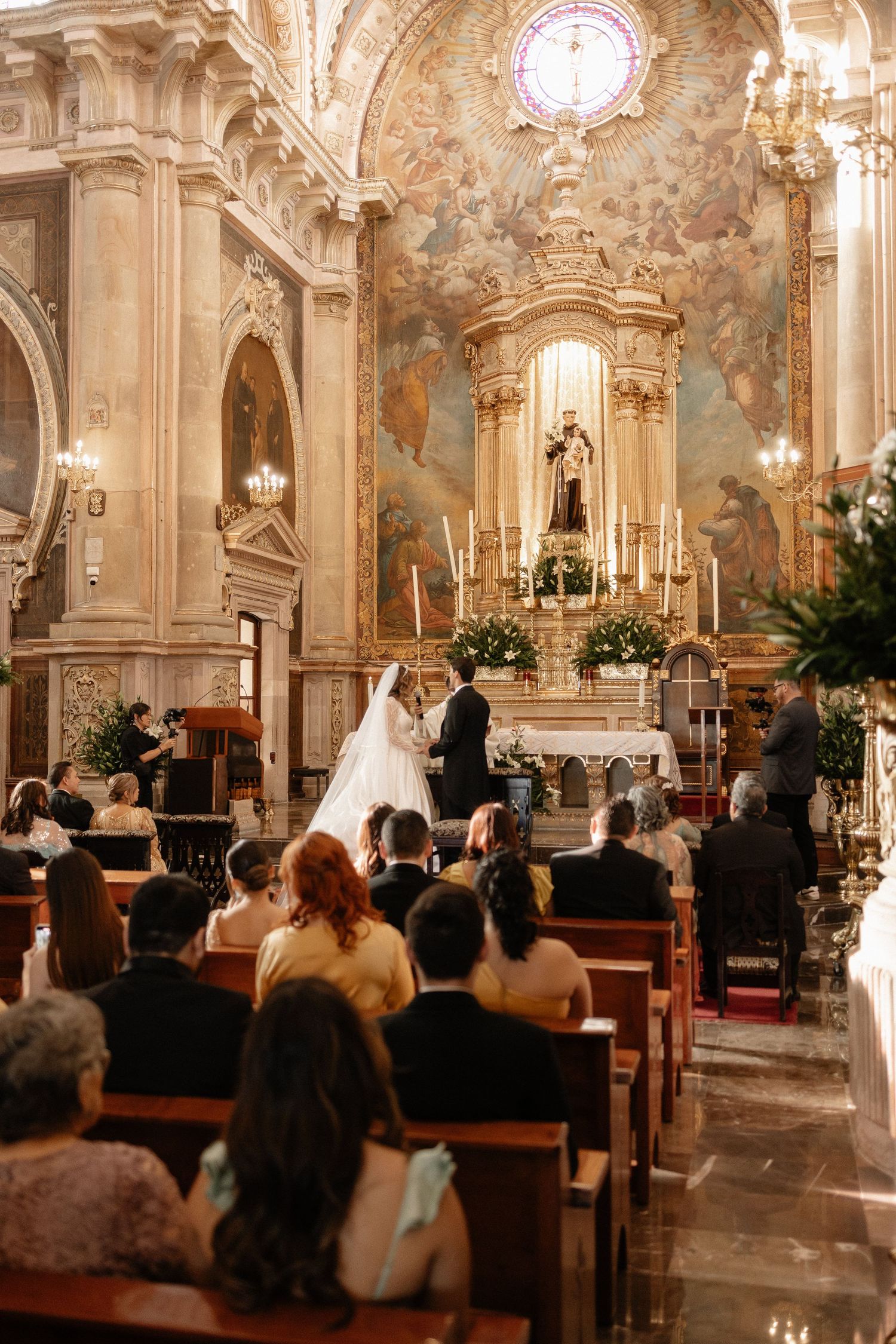 Wedding ceremony taking place inside an ornate Catholic church with elegant chandeliers and baroque architecture.