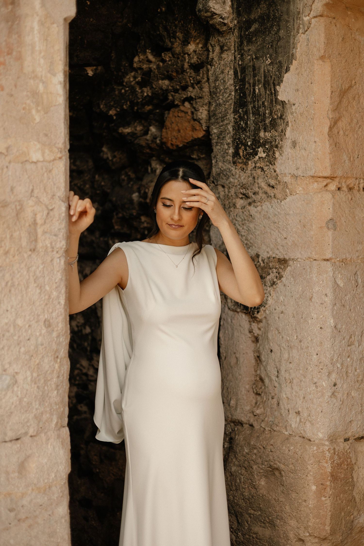 A person in a flowing white dress poses against a weathered stone wall in warm natural lighting.