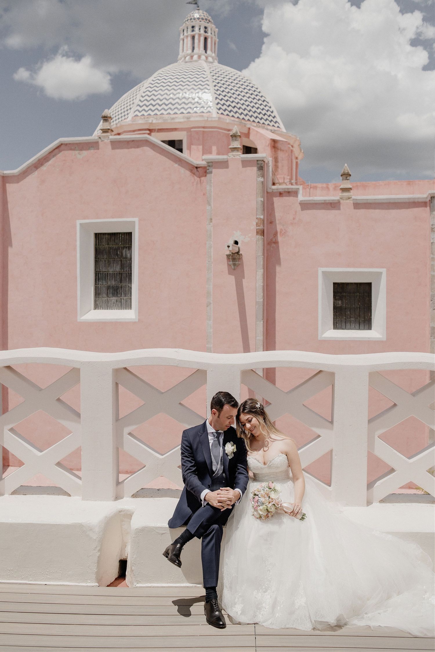 A couple poses romantically on a pink and white colonial church wall under cloudy skies.