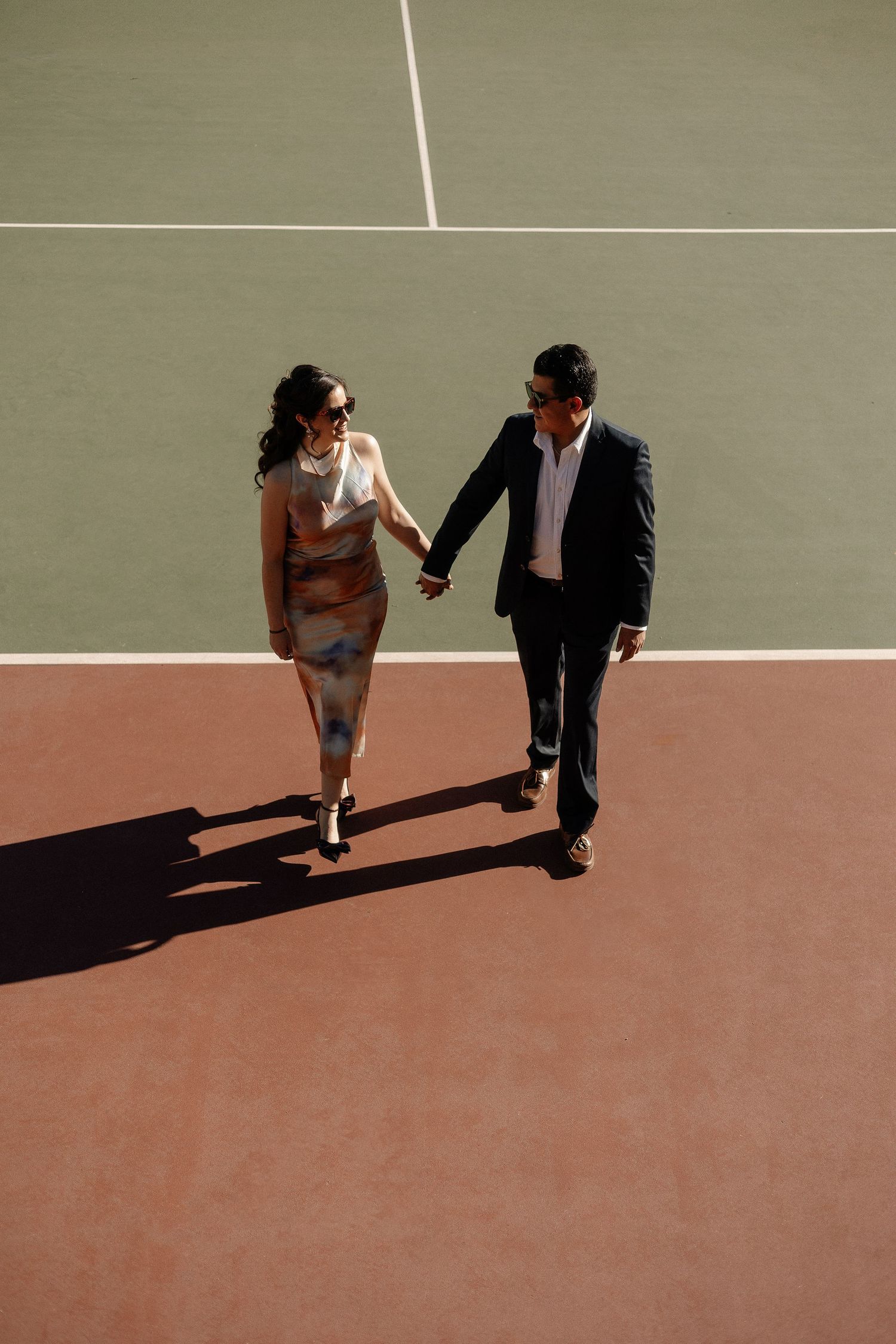 Two people in formal attire cast long shadows while walking on a tennis court at sunset.