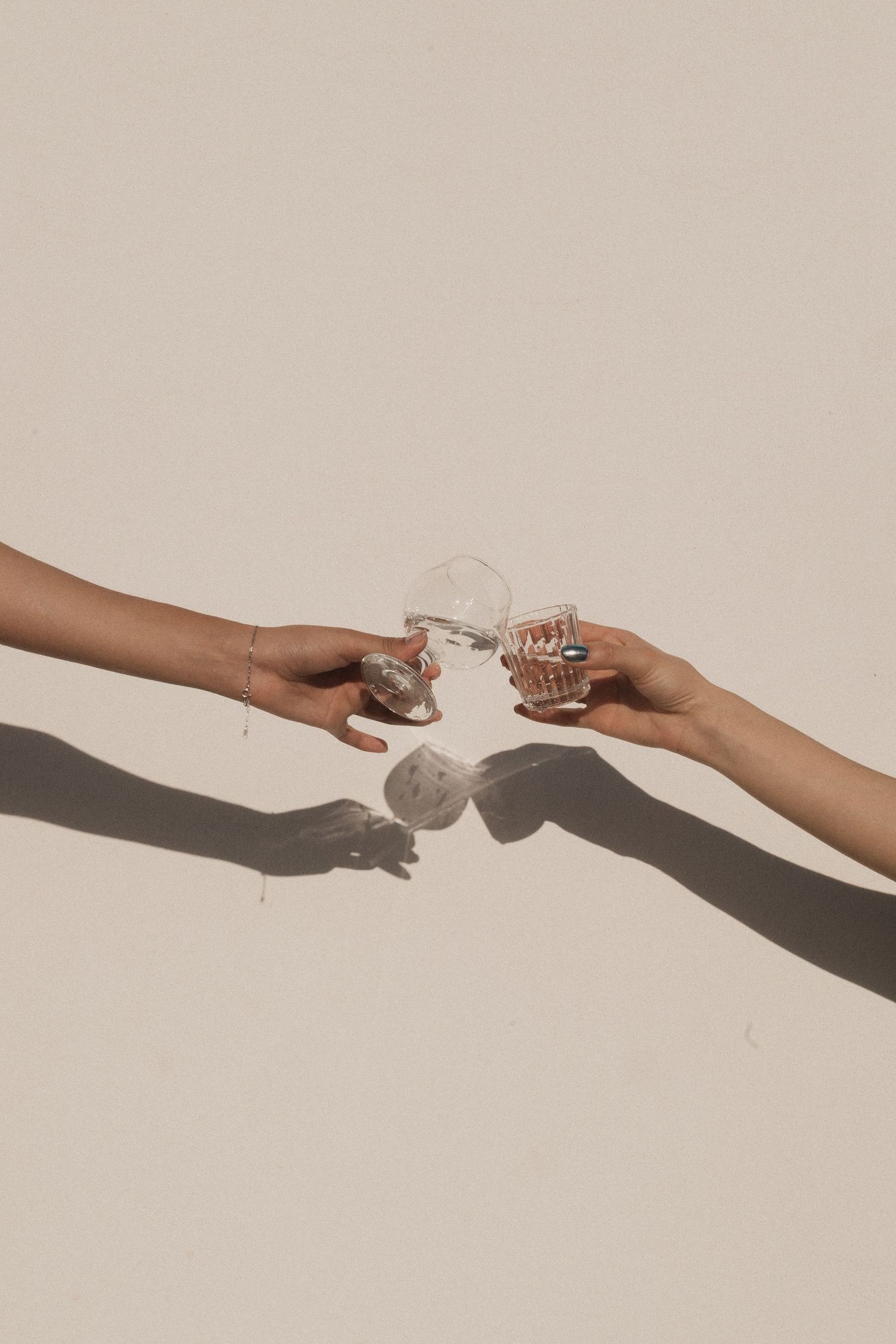 Two hands reaching to clink crystal glasses together against a beige background with dramatic shadows.