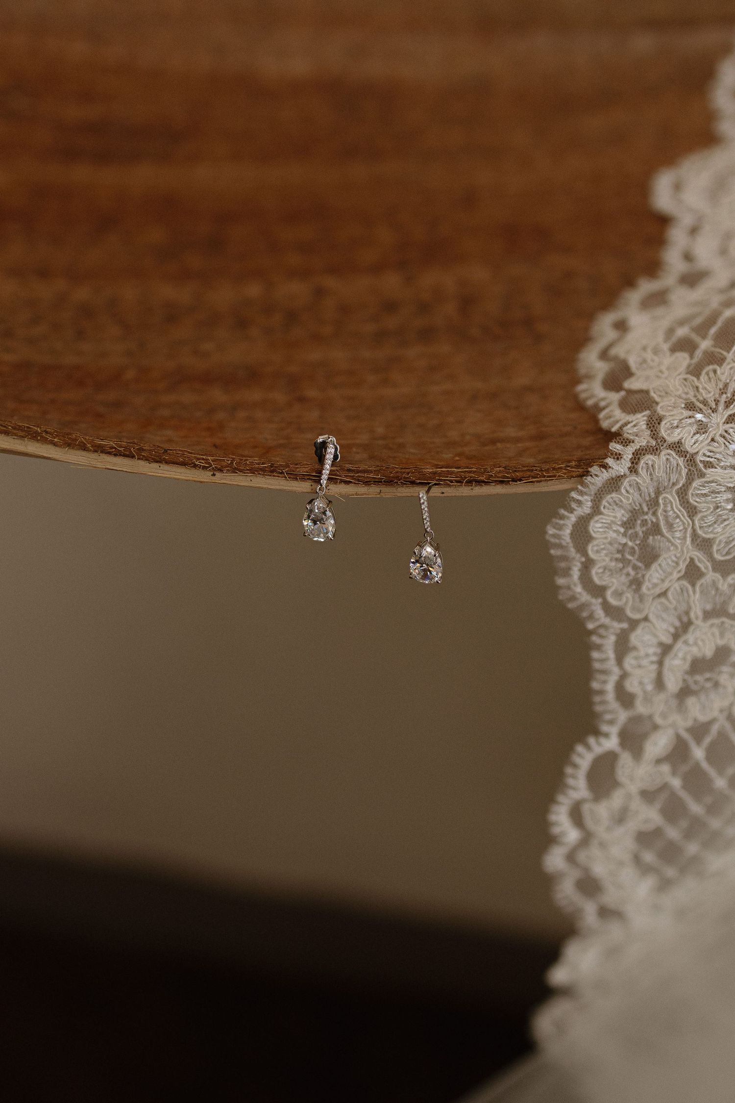 Delicate diamond stud earrings displayed next to white lace wedding dress detail on wooden surface.