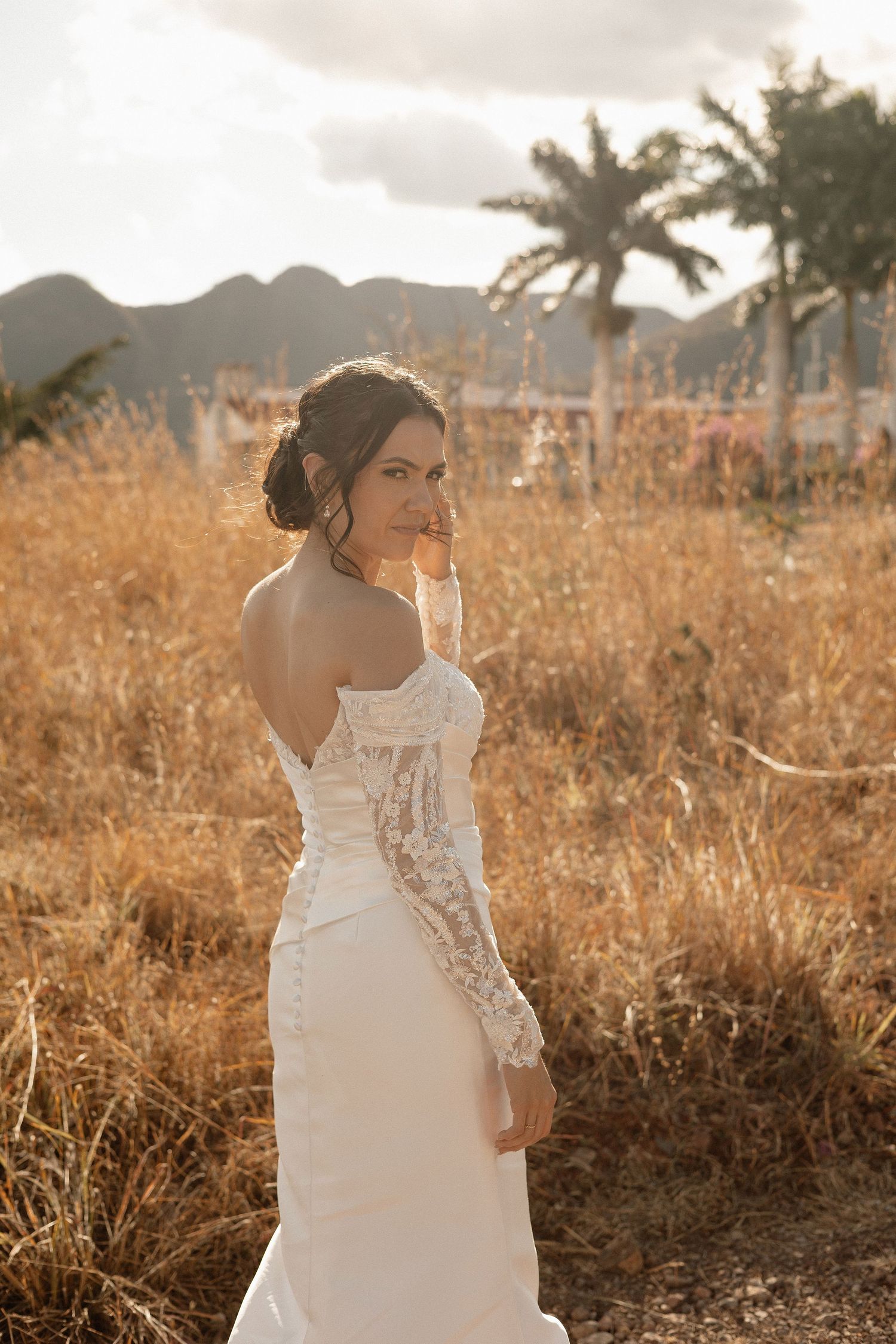 Bride in white lace dress poses in golden sunlight with palm trees and mountains in background at sunset.