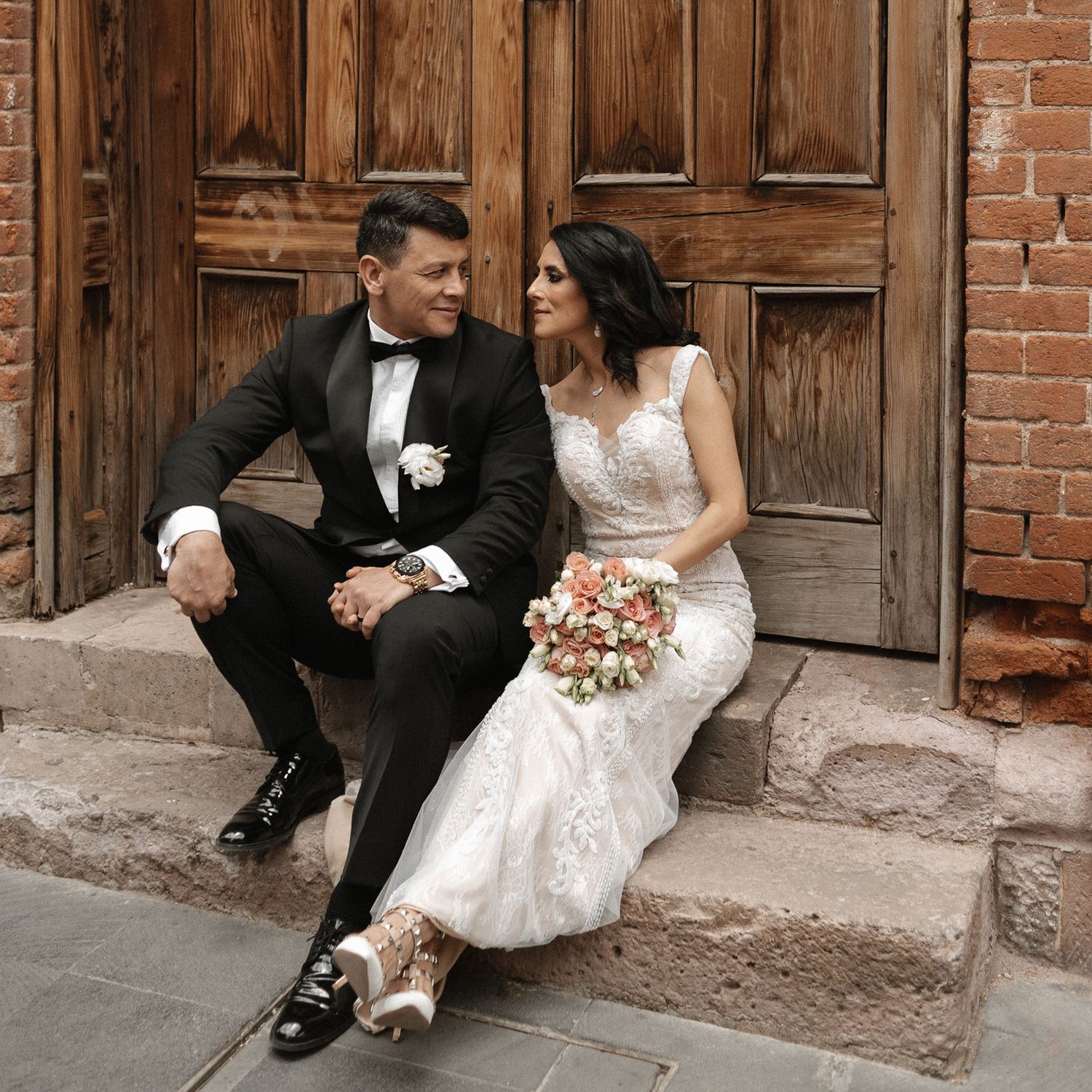 Wedding couple sitting casually on stone steps against rustic wooden doors.