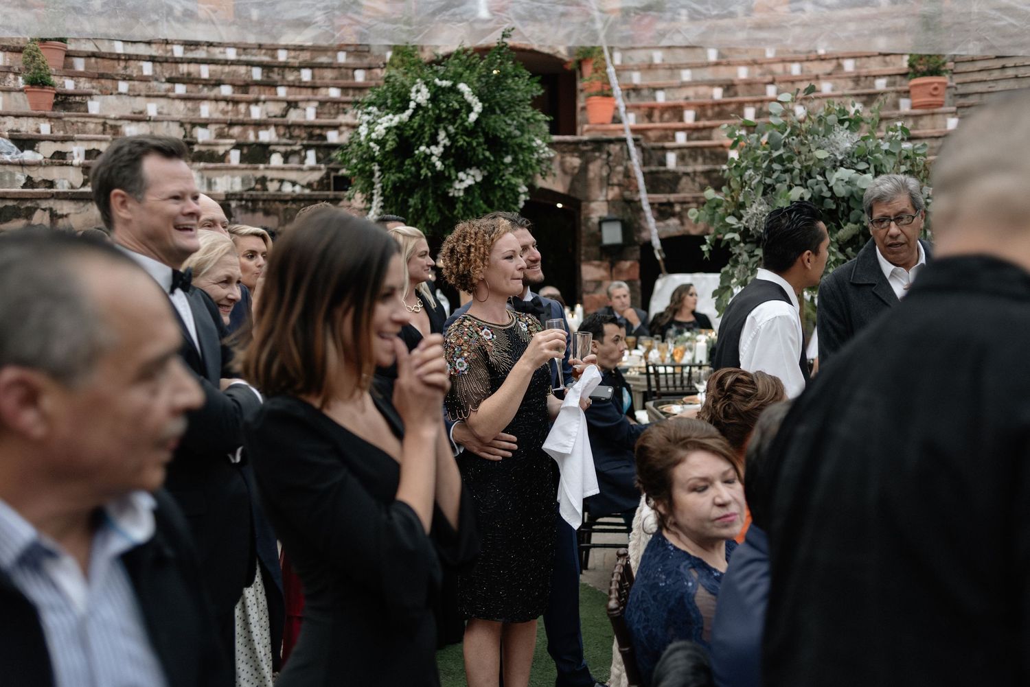 Wedding guests mingle and socialize in an outdoor courtyard setting with exposed brick walls and hanging greenery.