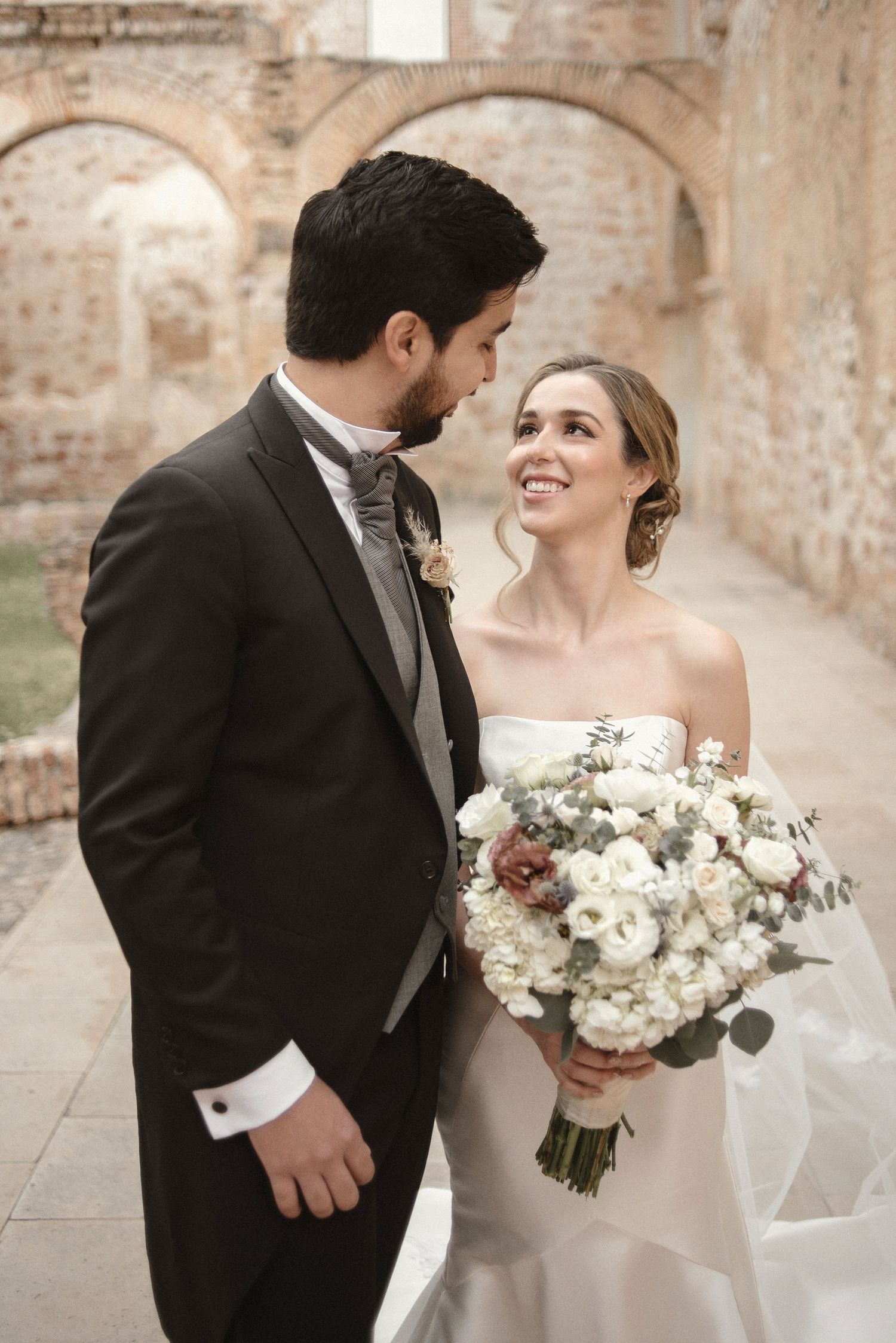 A tender moment captured between a couple with a white floral bouquet against stone archways.
