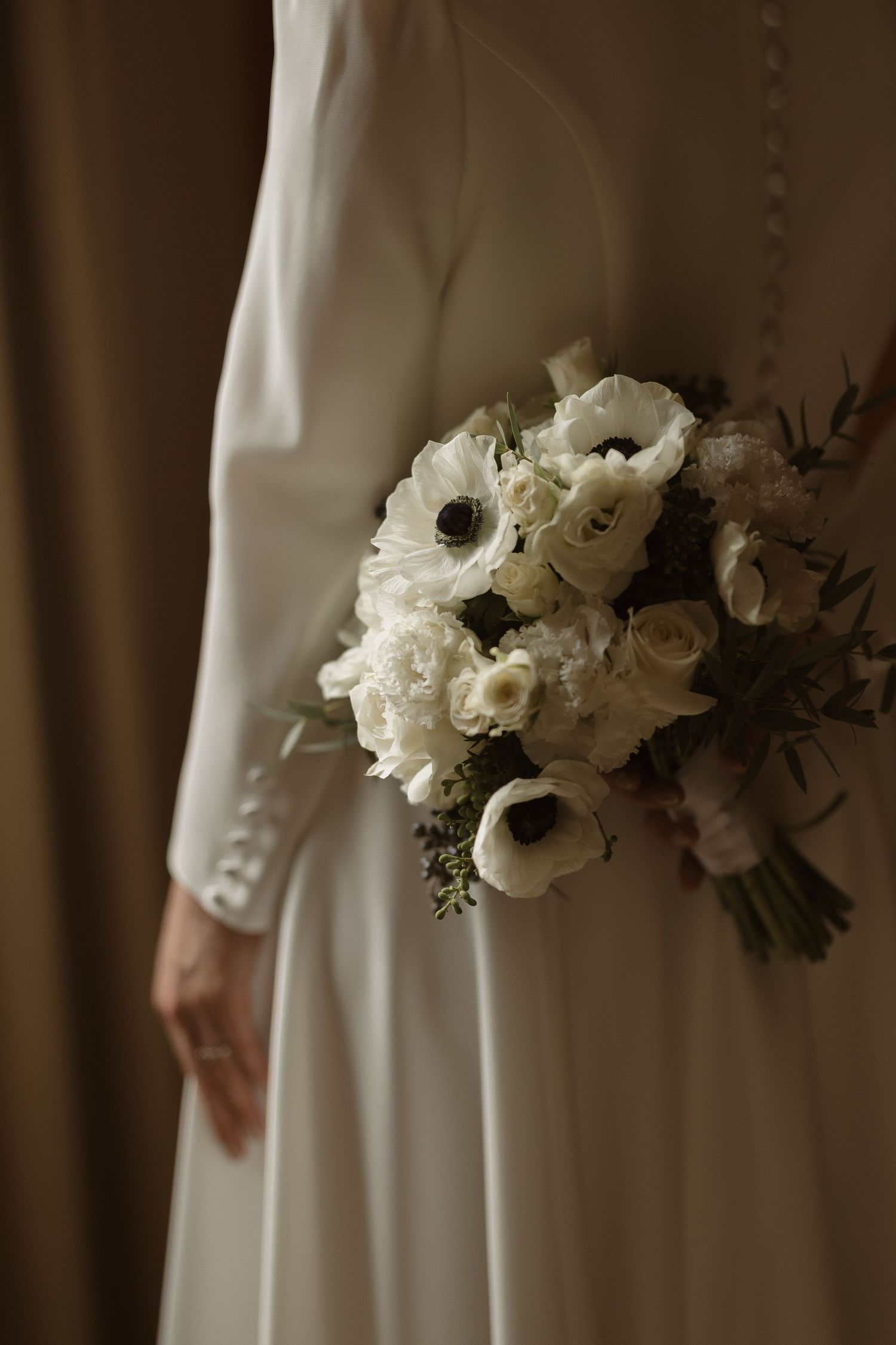 A delicate white wedding bouquet featuring anemones and ranunculus flowers against a flowing white dress.