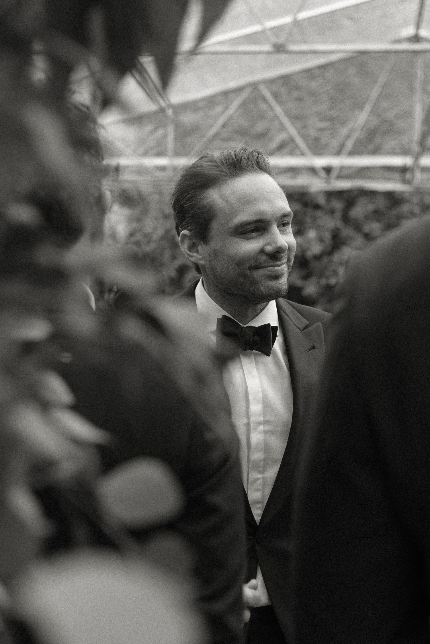 Black and white formal portrait of a smiling person in a classic tuxedo with bow tie framed by blurred foliage.