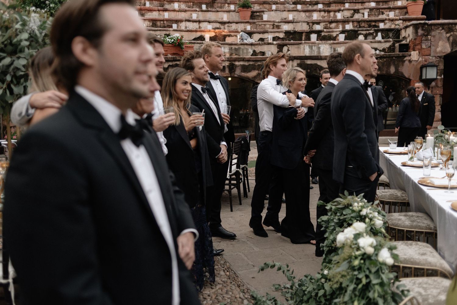 Wedding guests in black tie attire socialize at an outdoor reception near a brick wall and greenery.