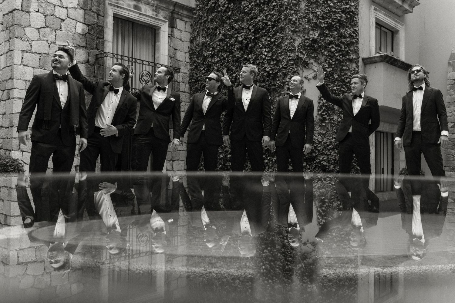Group of people dressed in vintage formal attire with bow ties and cuffed pants standing in a row against a stone building.